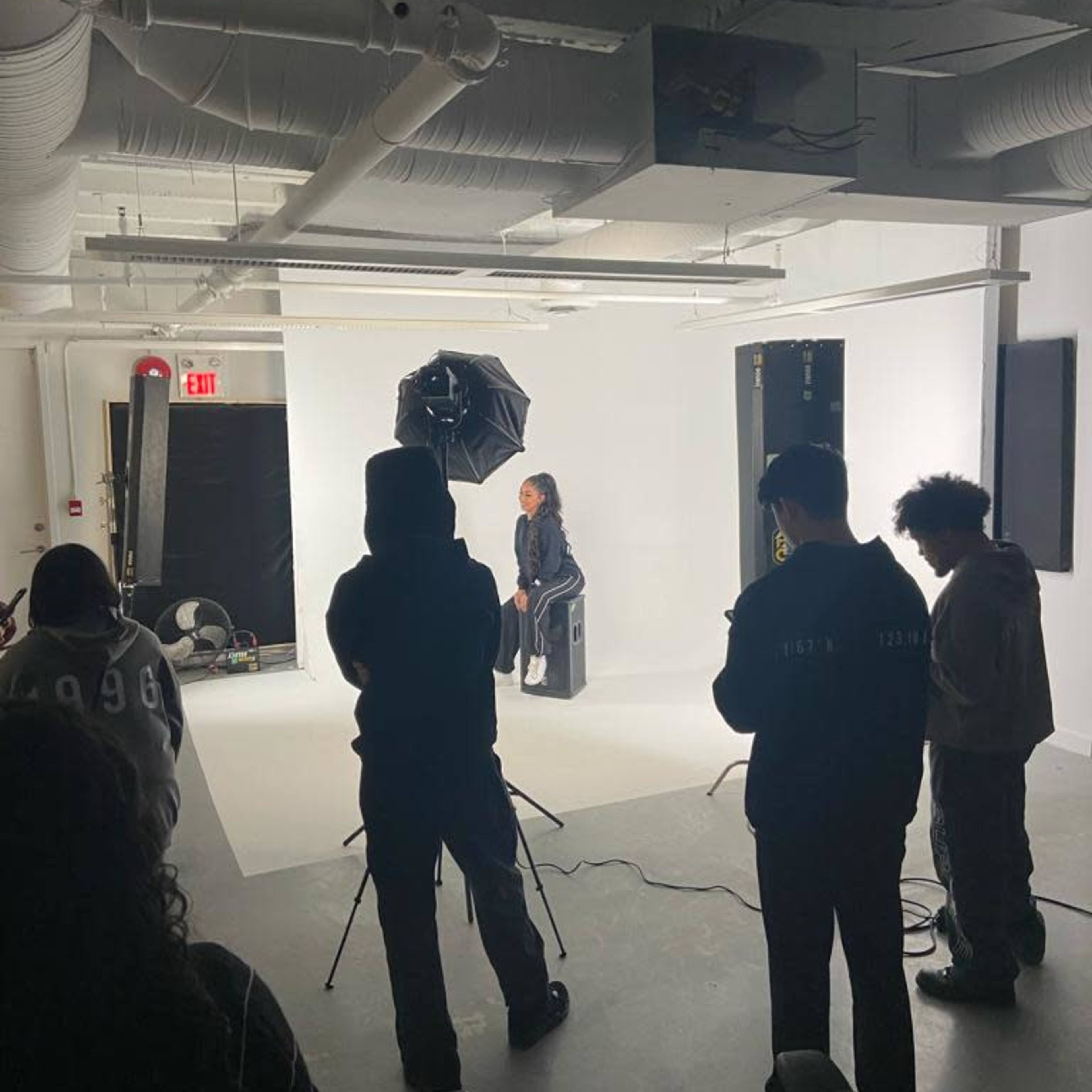 A photography session is taking place in a studio with a seated model and several people observing and adjusting equipment.