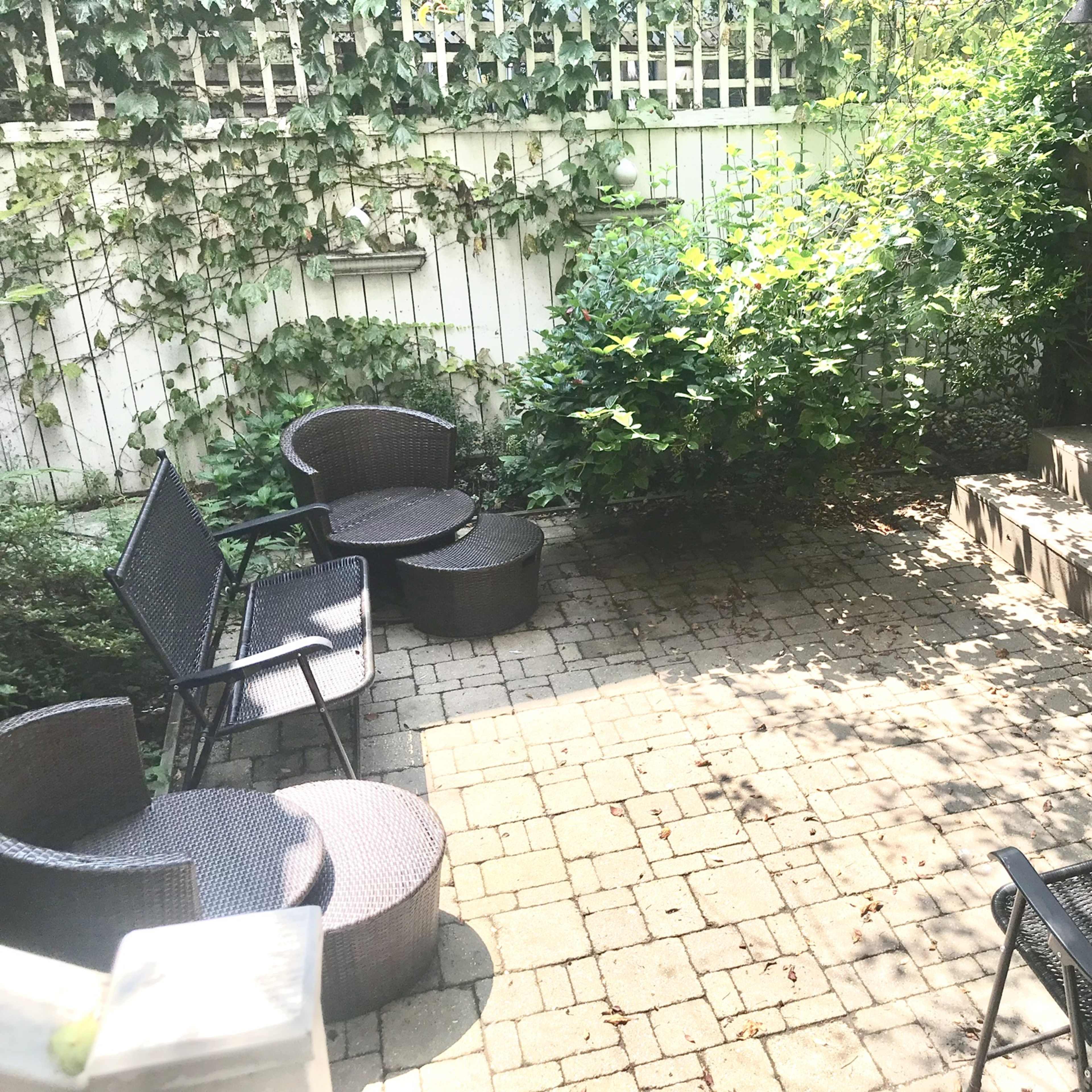 A small outdoor patio features several wicker chairs and tables arranged around a green bush, with a wooden fence and a blue door in the background.