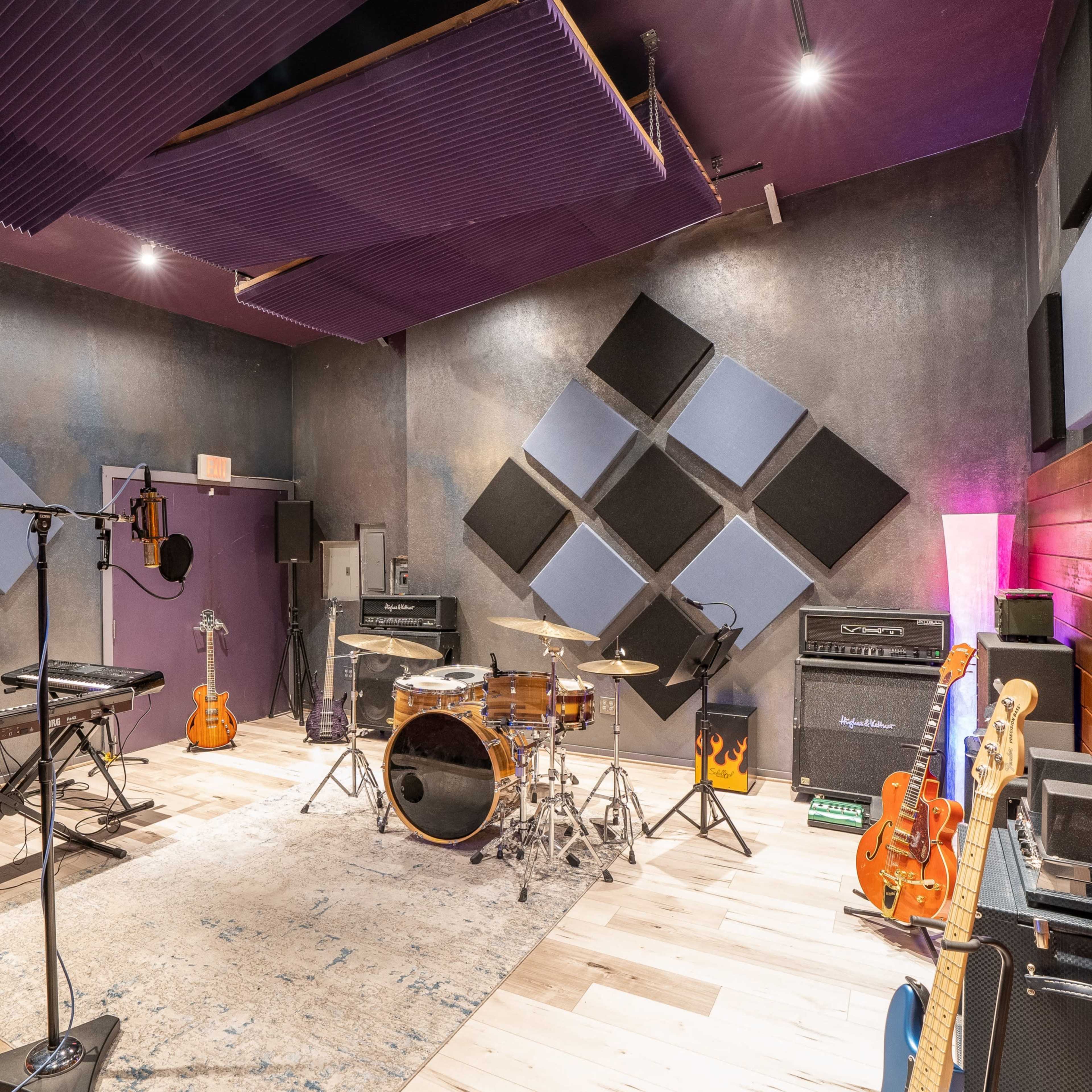 A music recording studio featuring a drum set, keyboard, guitar amplifiers, and various instruments arranged around a rug on a wooden floor.