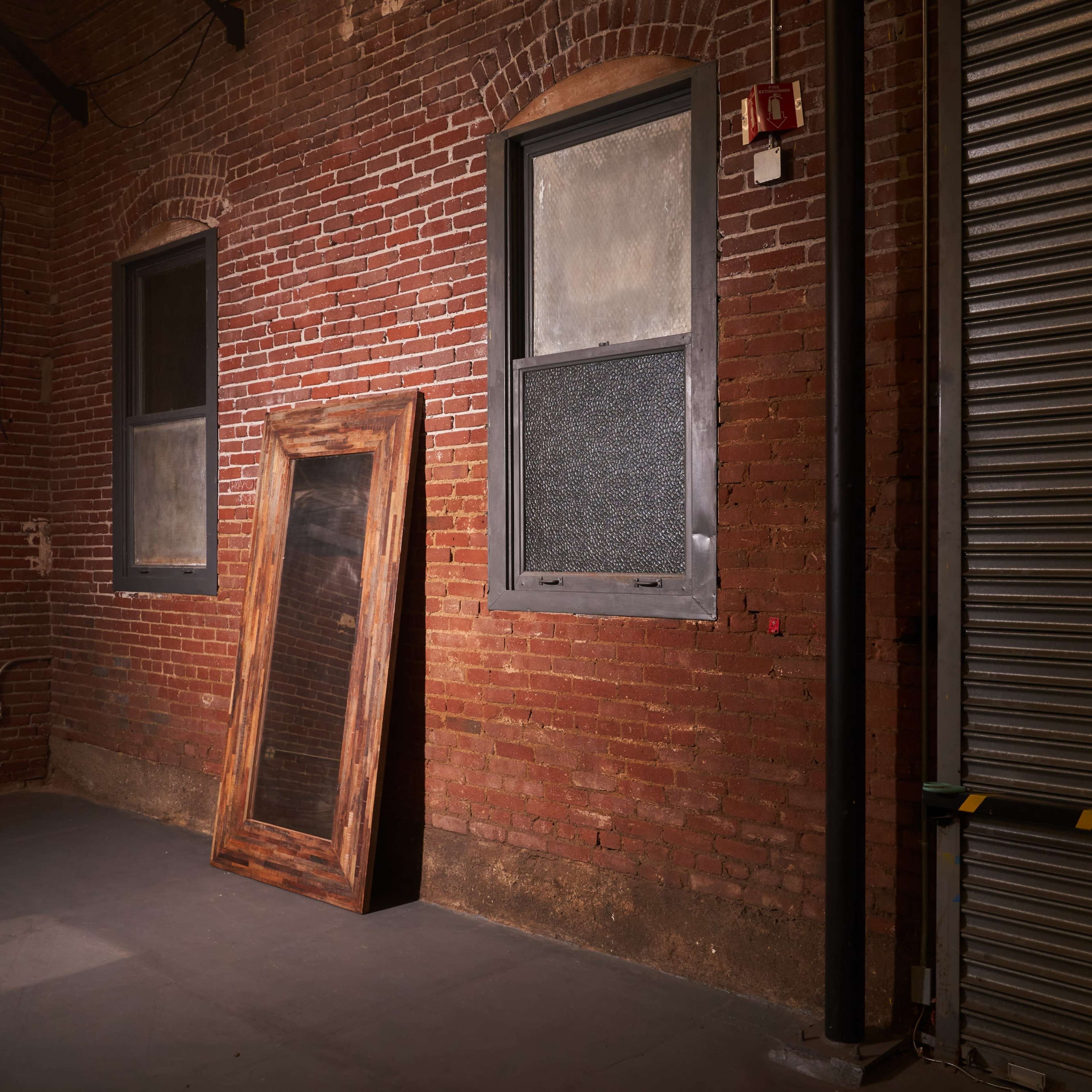 A room with exposed brick walls, featuring a large wooden-framed mirror leaning against the wall next to a metal roll-up door.