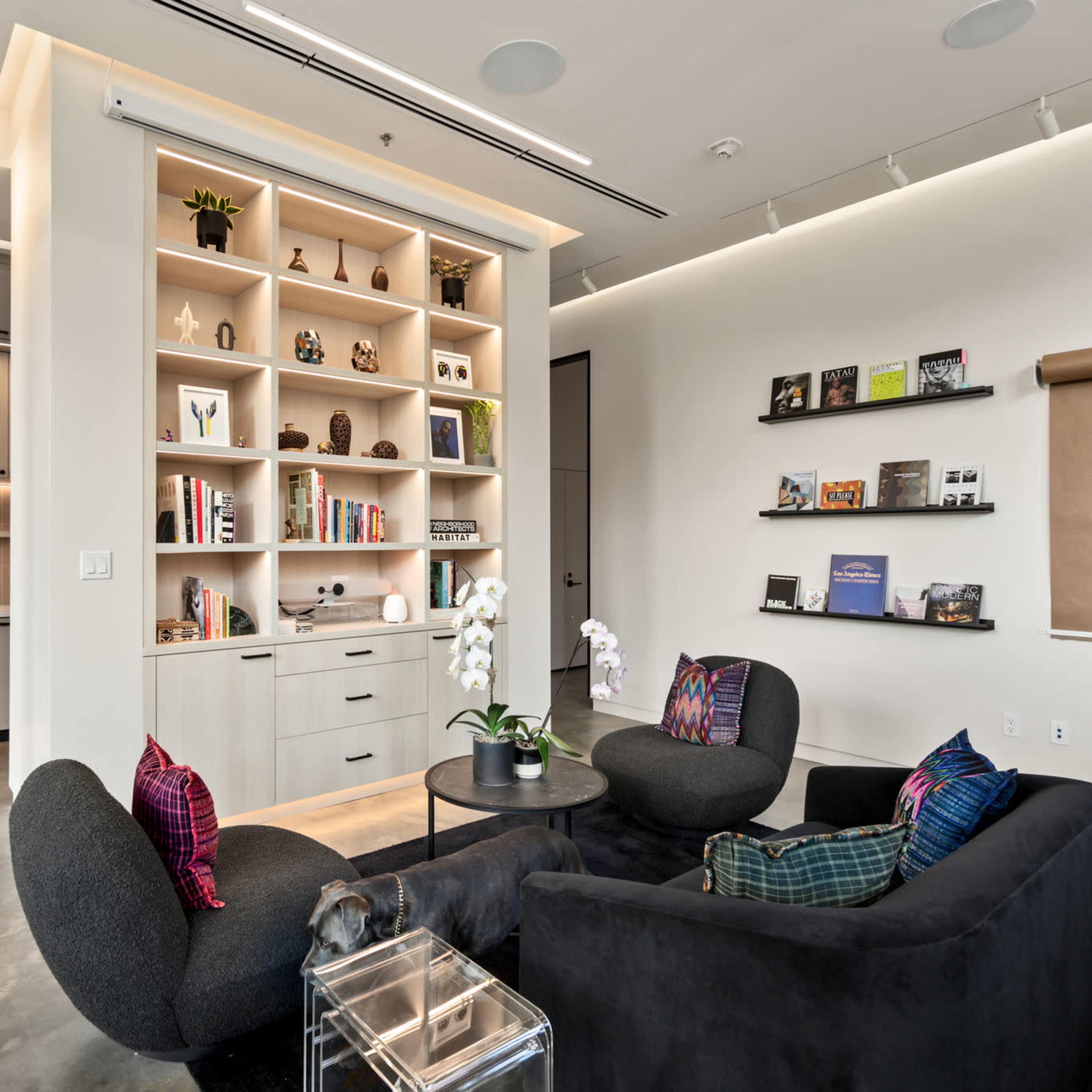 A modern living area features two black chairs, a circular table, and a built-in shelving unit displaying books and decorative items.