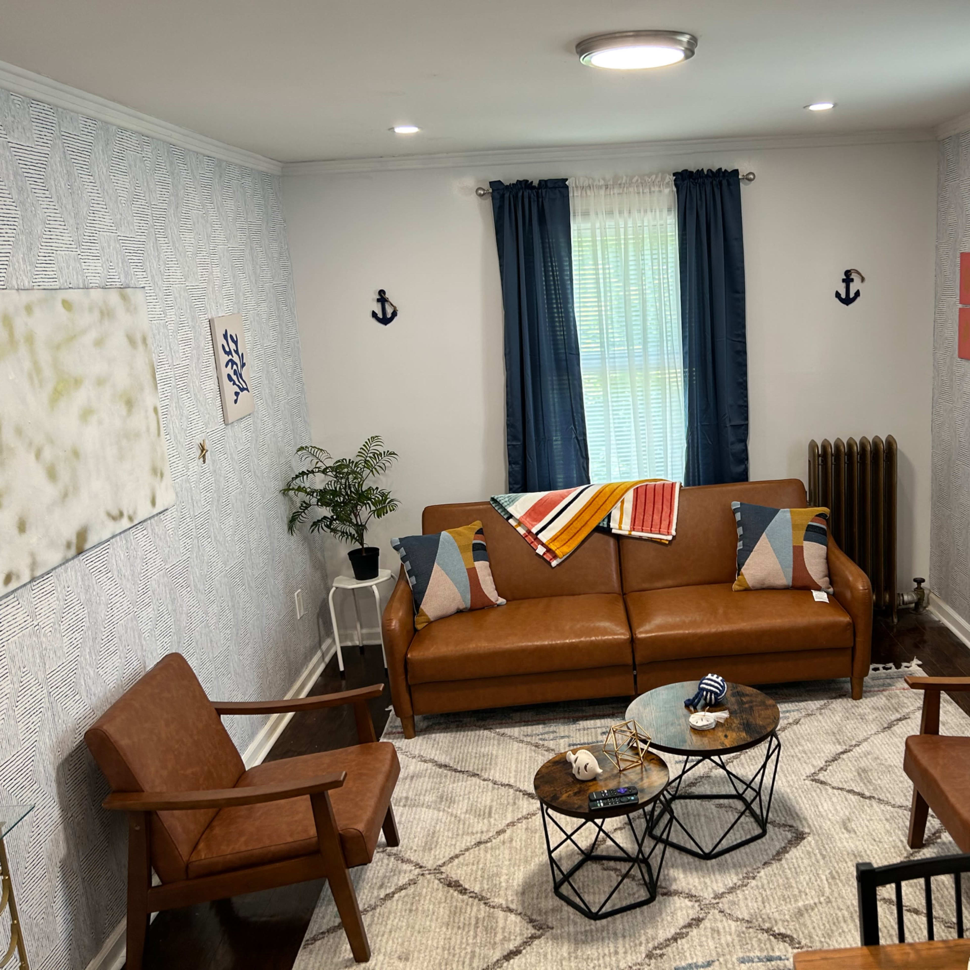 A cozy living room with a brown leather sofa, two tan armchairs, a circular coffee table, and nautical-themed decor against patterned wallpaper.