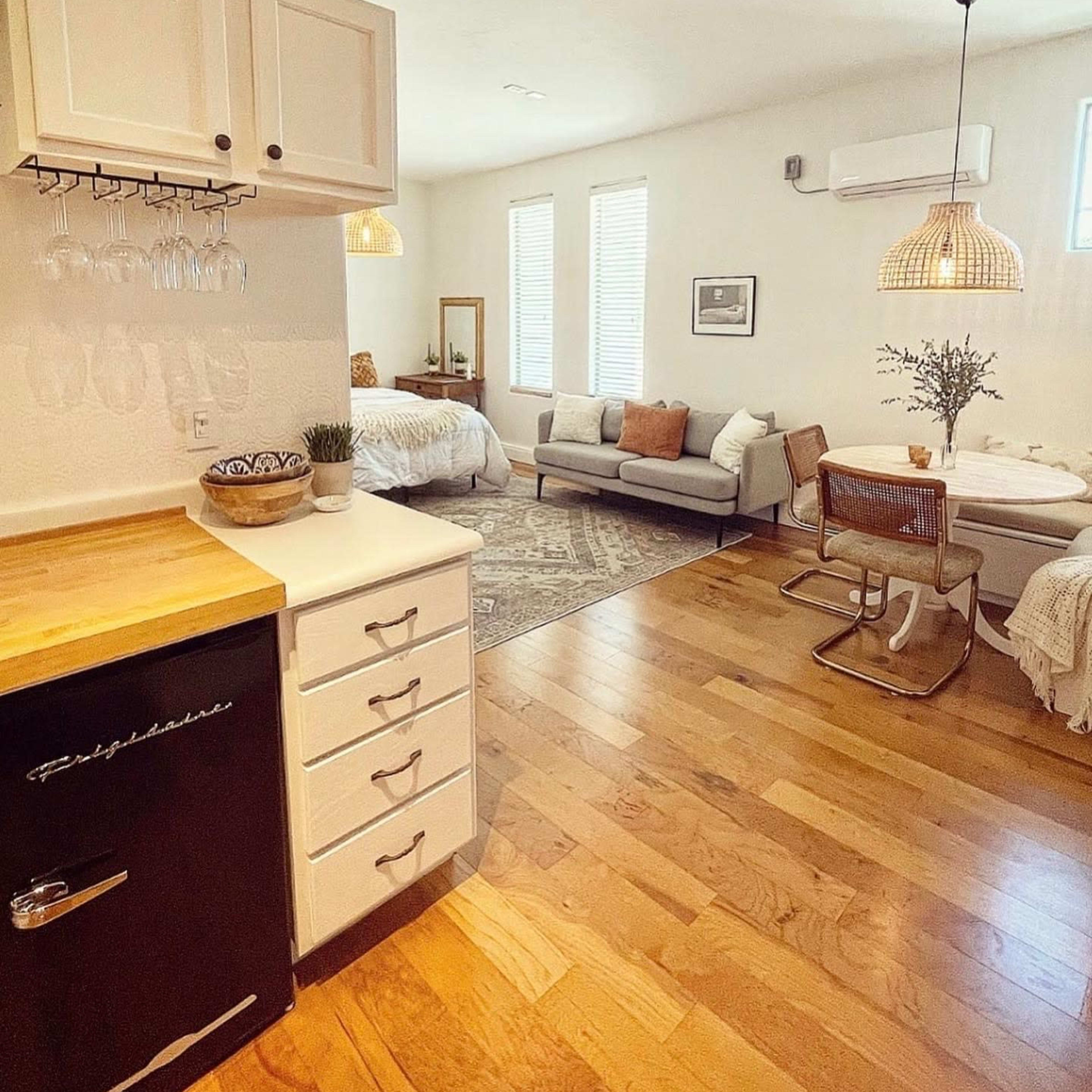 A small, open-concept living space features a kitchenette, seating area, and a bedroom with natural light streaming through windows.