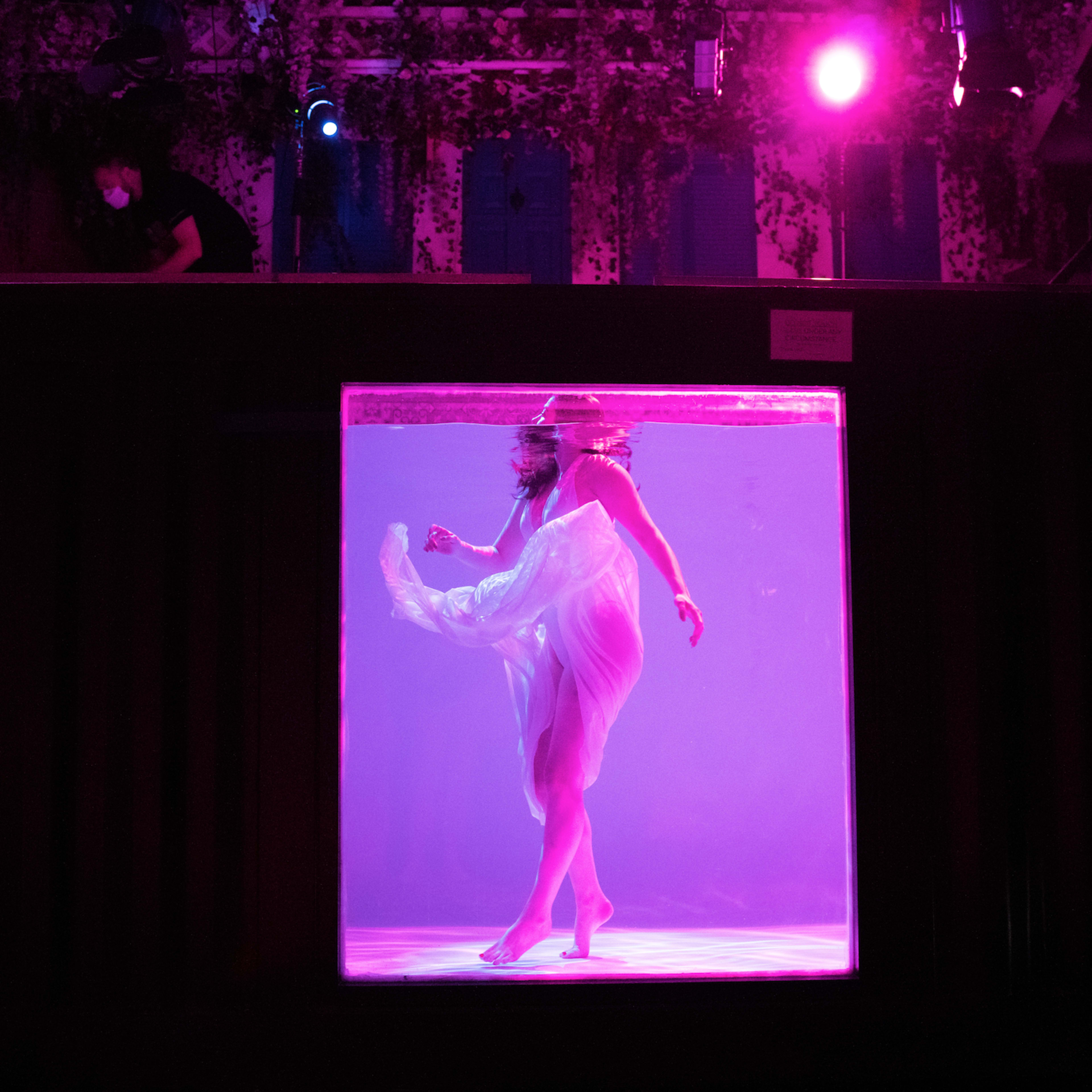 A performer dances in a submerged glass enclosure while a person operates equipment above in a dimly lit venue.