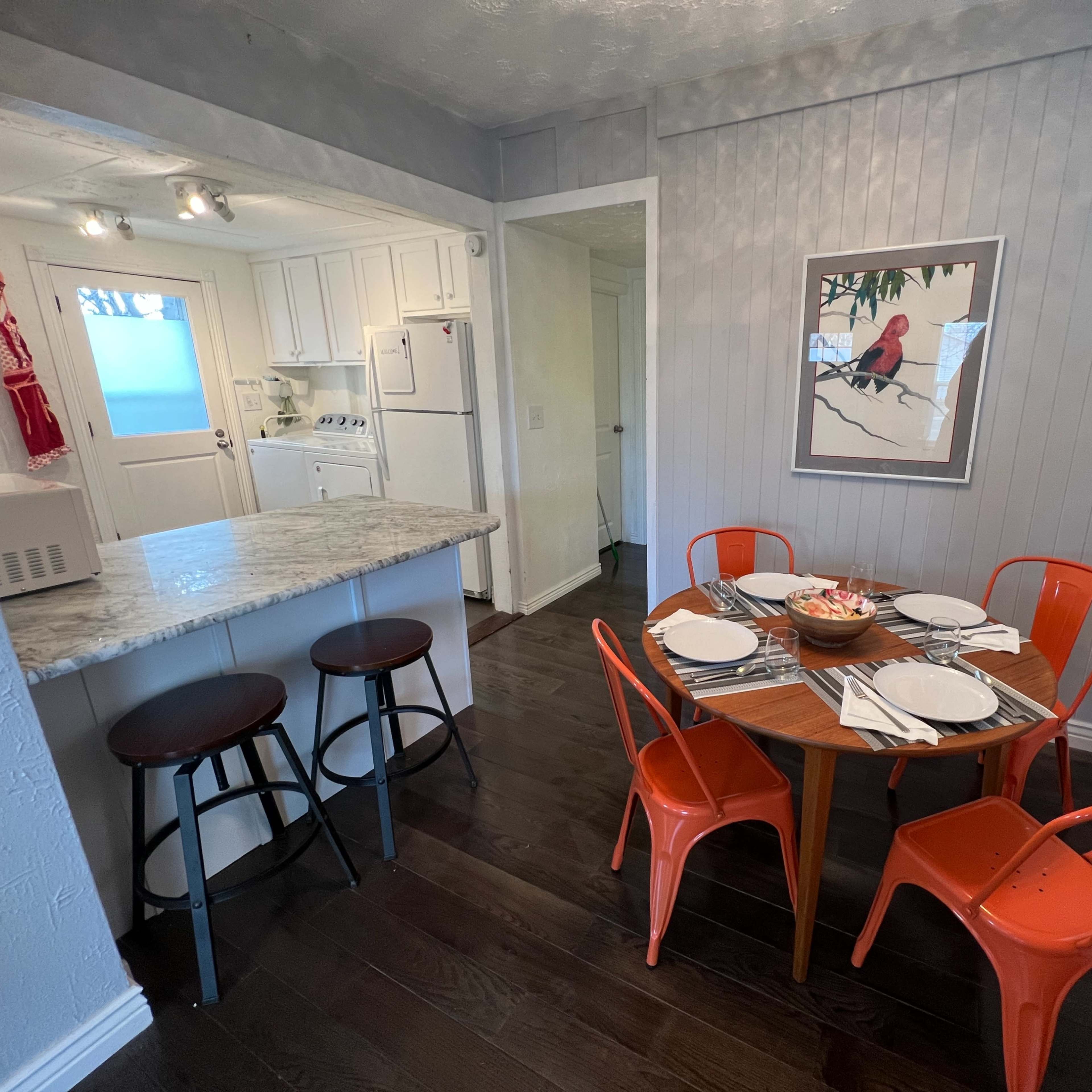 A small dining area features a round wooden table set for four, surrounded by orange metal chairs, with a view of a kitchen area in the background.