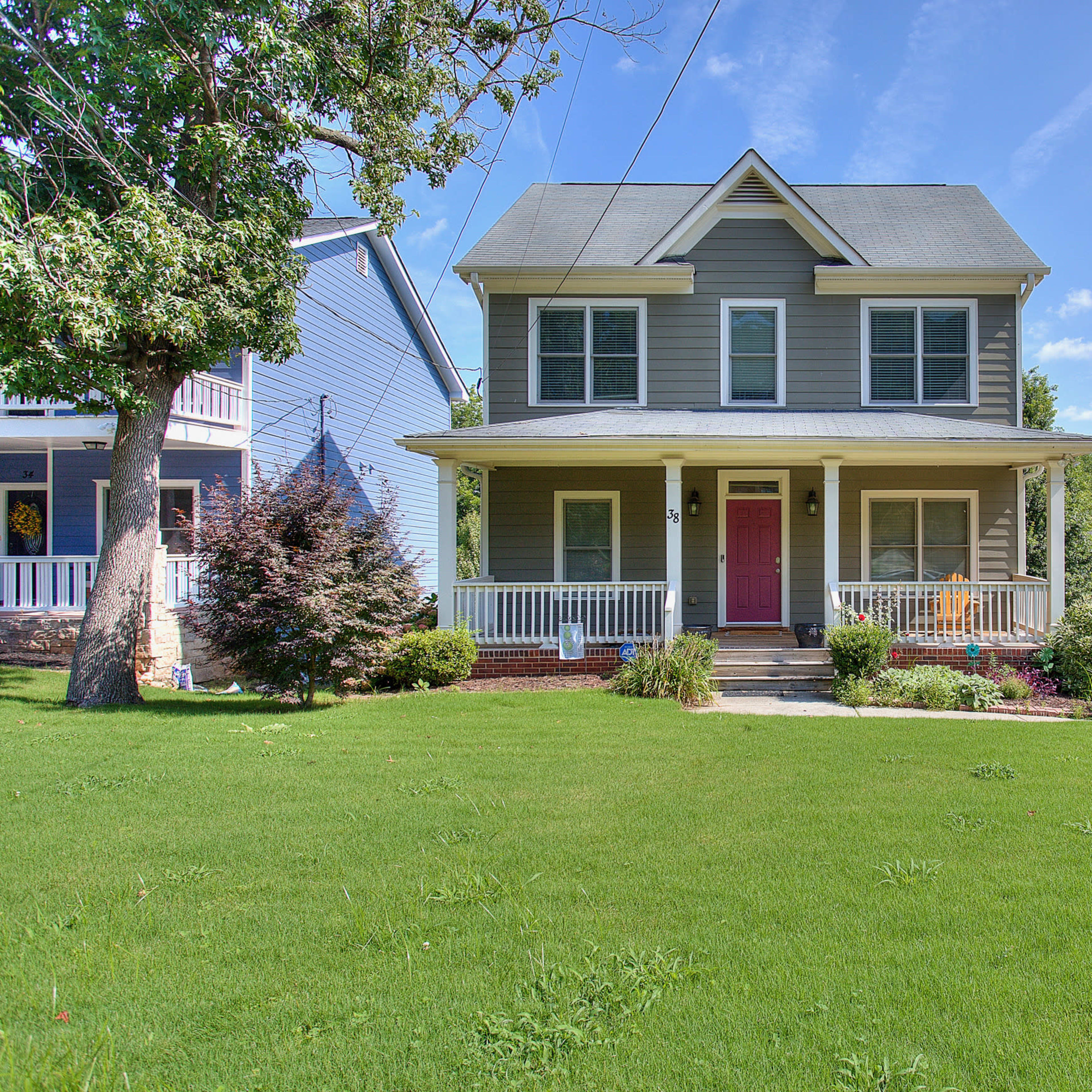 A two-story gray house with a front porch and a red door is surrounded by a green lawn and shrubbery.