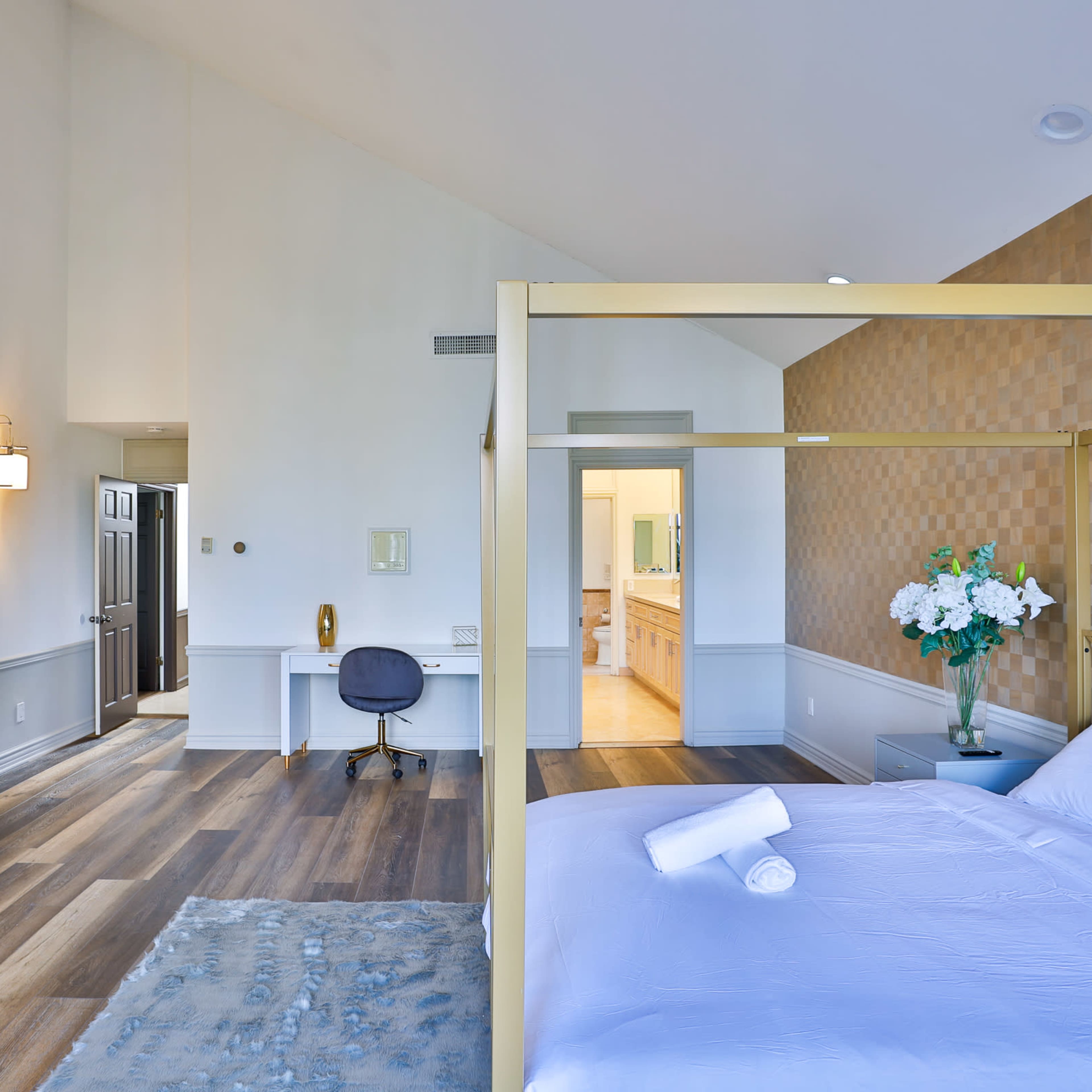A modern bedroom features a four-poster bed, a fireplace, a desk area, and an en-suite bathroom beyond a doorway.