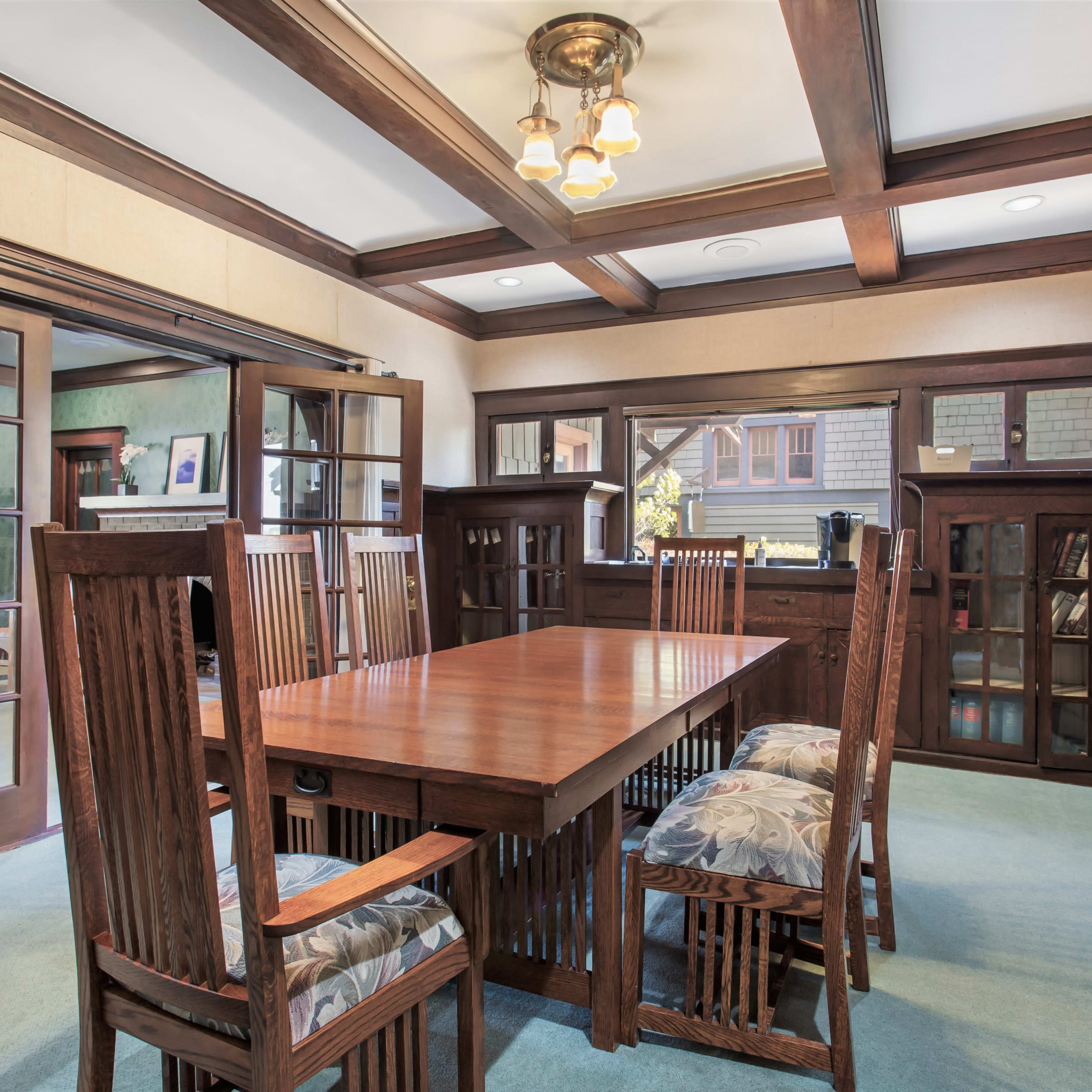 A wooden dining room with a large table surrounded by six chairs, large windows allowing natural light, and built-in shelves against the walls.
