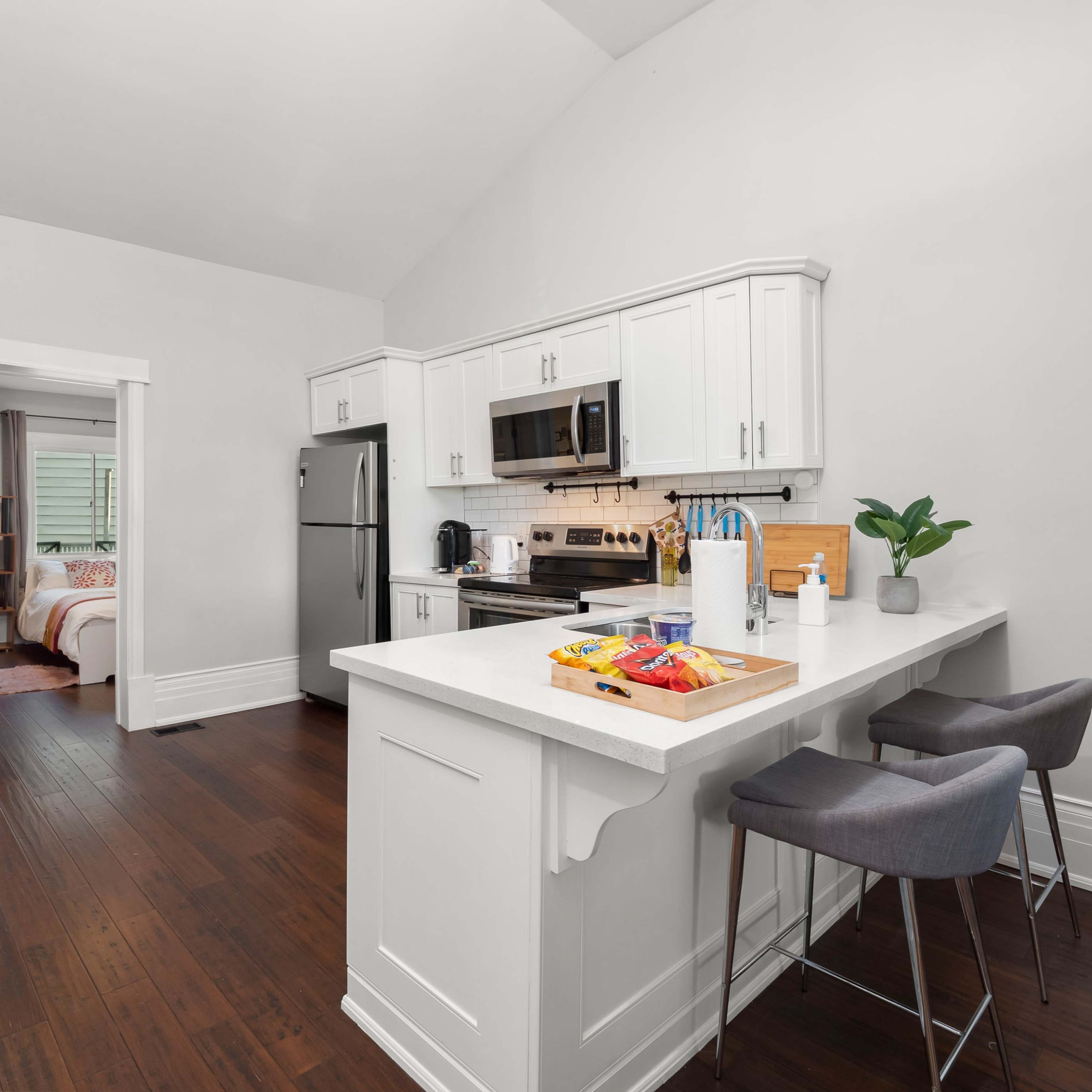 A modern kitchen area with white cabinetry, a stainless steel refrigerator, and a bar counter with two stools, opening up to a living space.