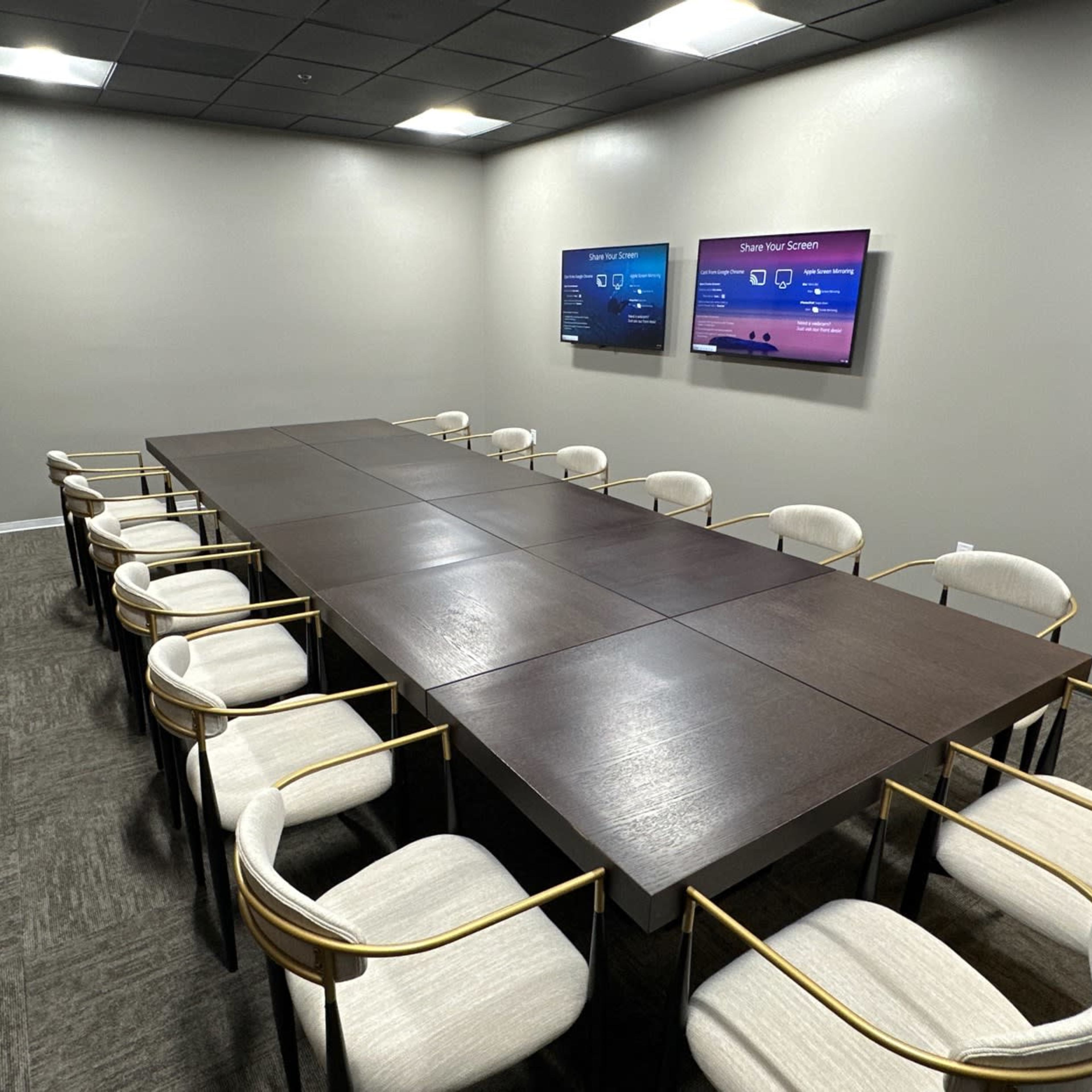 A modern conference room with a long wooden table surrounded by twelve chairs and two screens mounted on the wall.