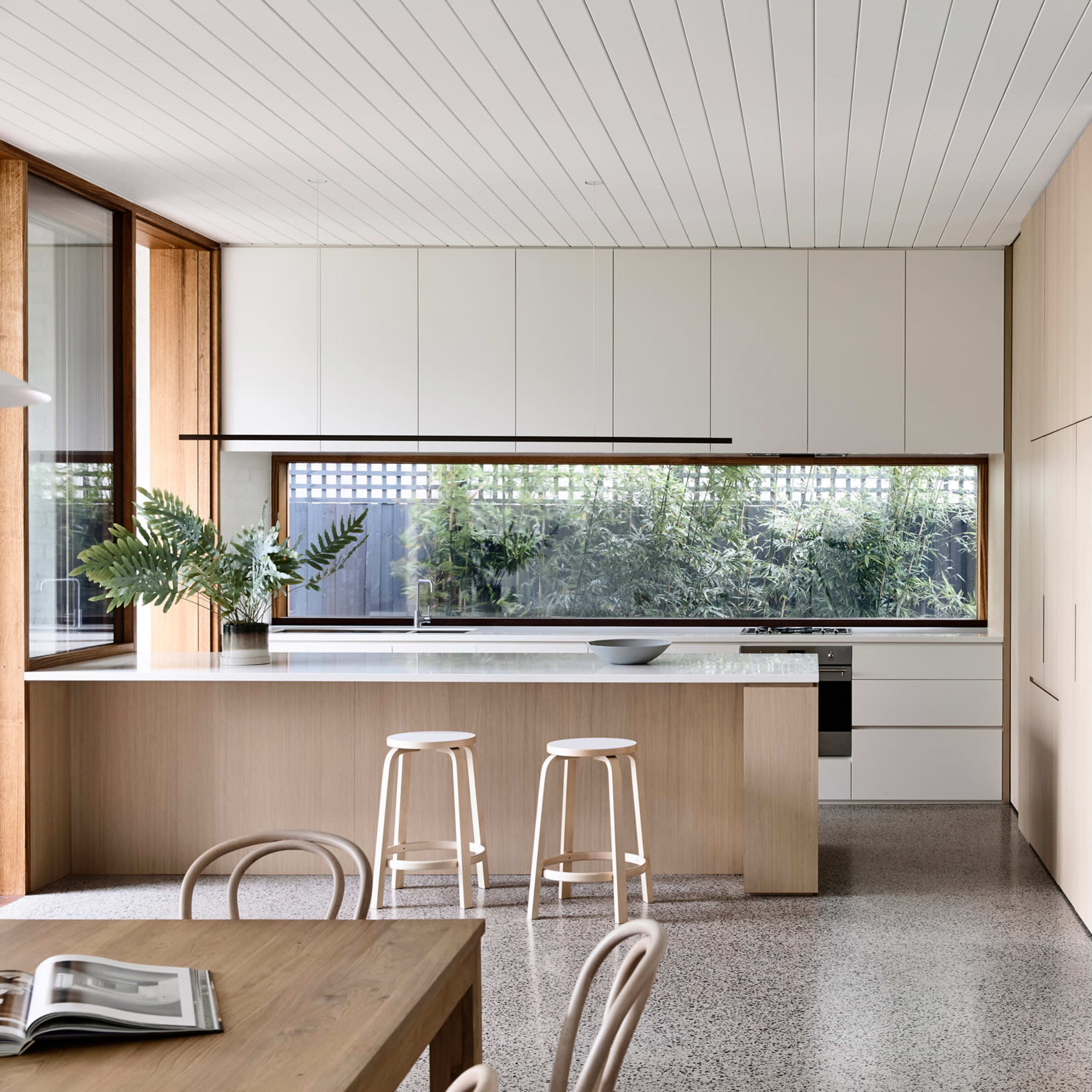 A modern kitchen with light wood cabinetry, a central island with three stools, and large windows overlooking a garden.