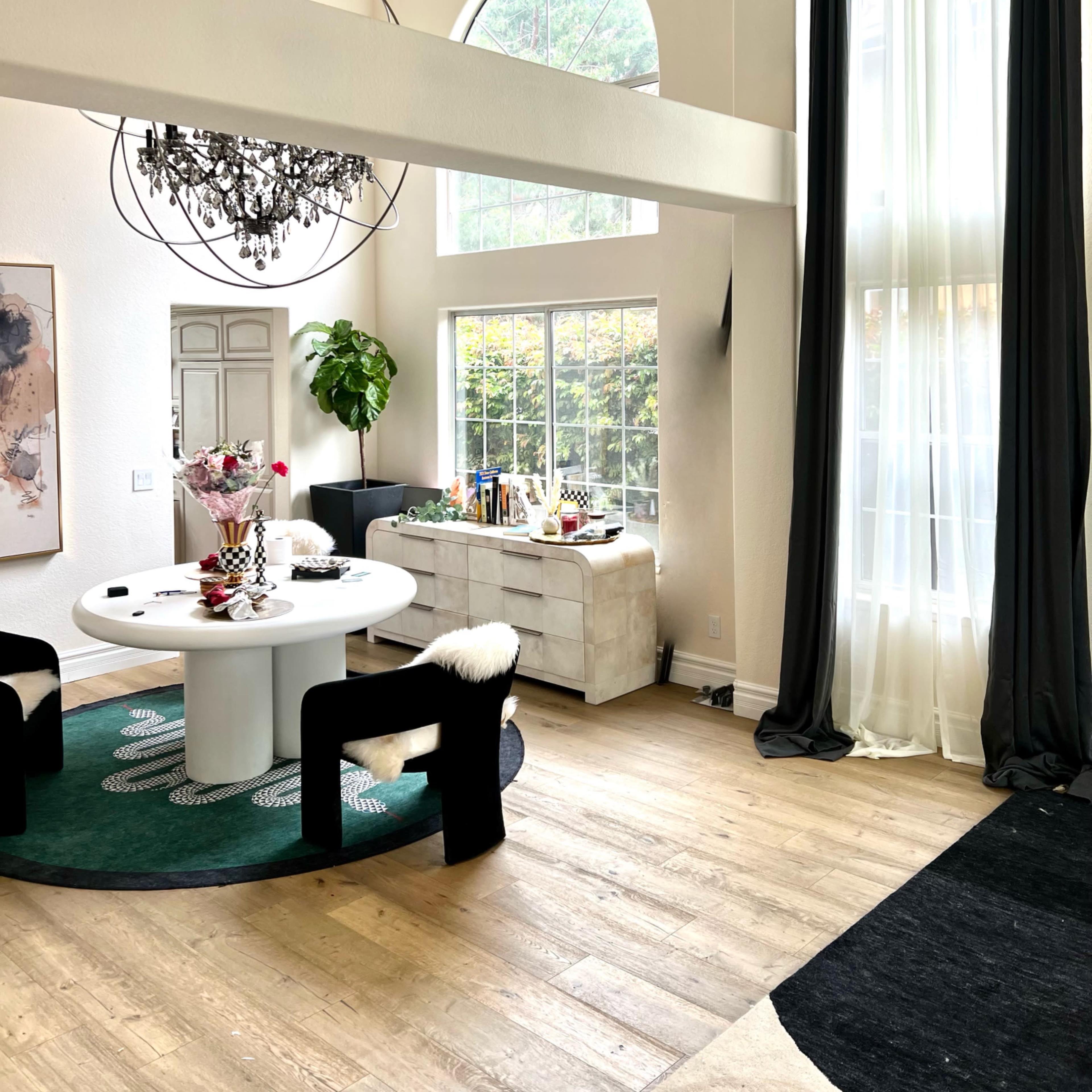 A modern dining area featuring a round table surrounded by black chairs, a large window with sheer curtains, and a chandelier hanging from the ceiling.