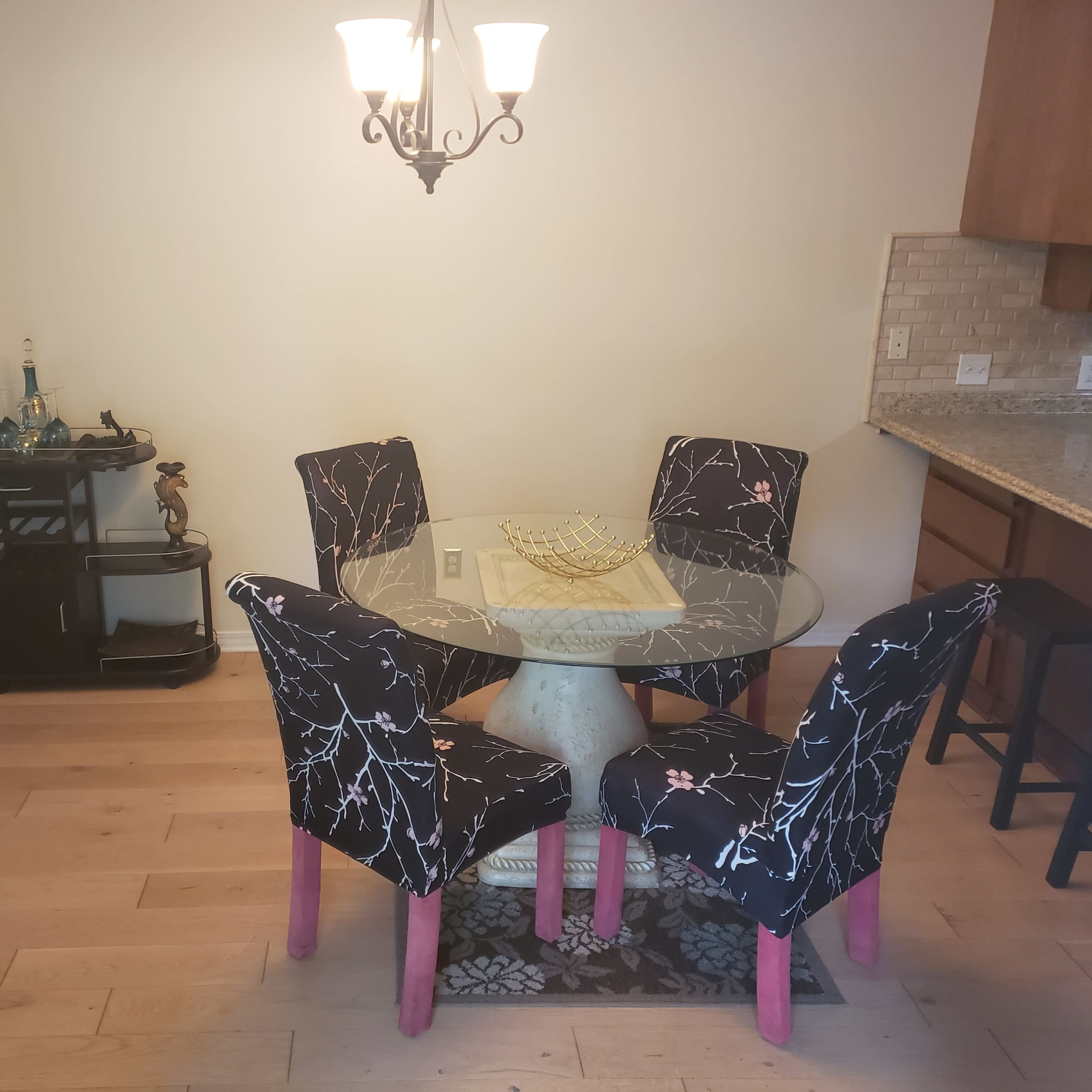 A glass-top dining table with four patterned chairs is positioned in a light-colored room alongside a small bar cart and lamp.