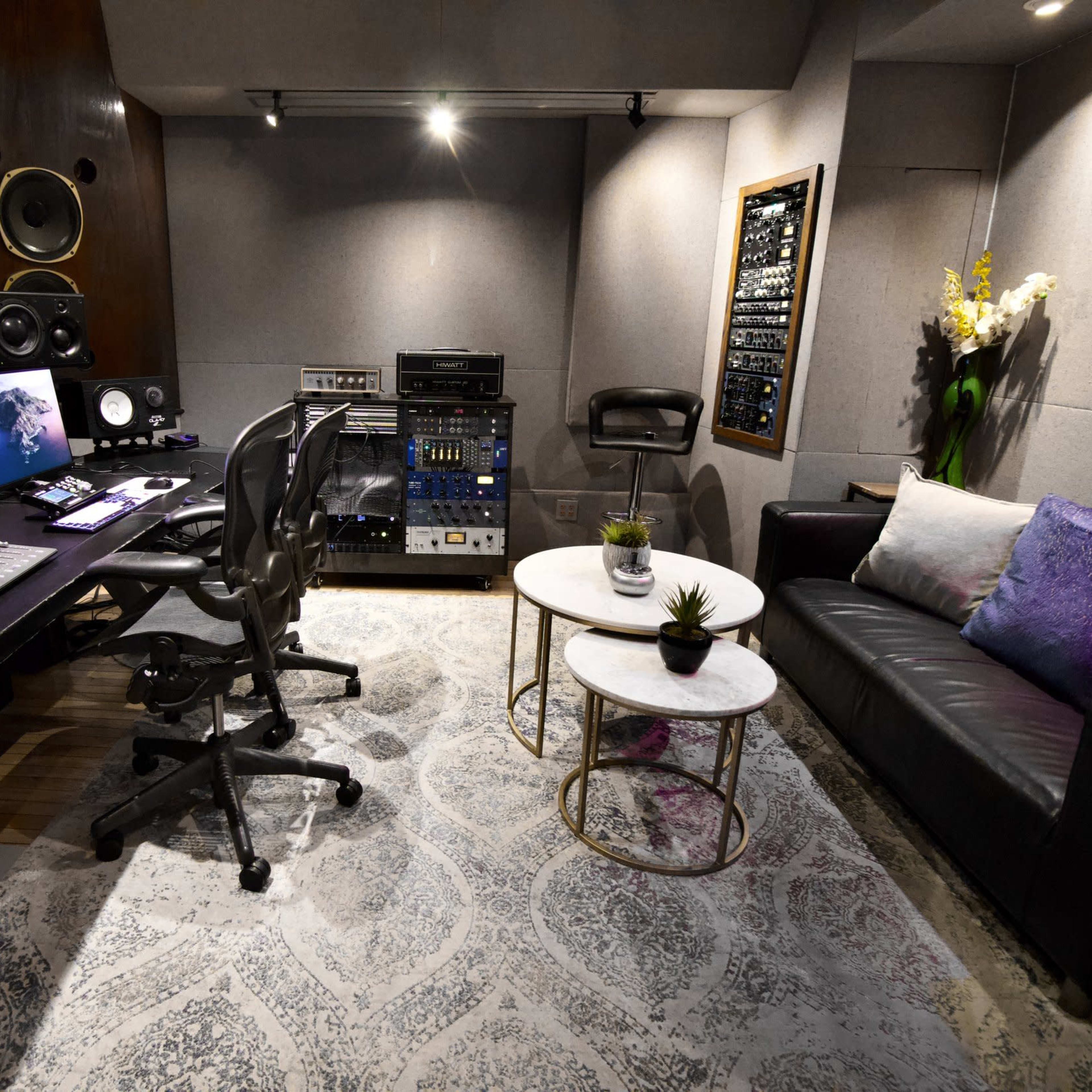 A modern music studio featuring dual computer monitors, a control desk with mixing equipment, a black sofa, and a round coffee table on a patterned rug.