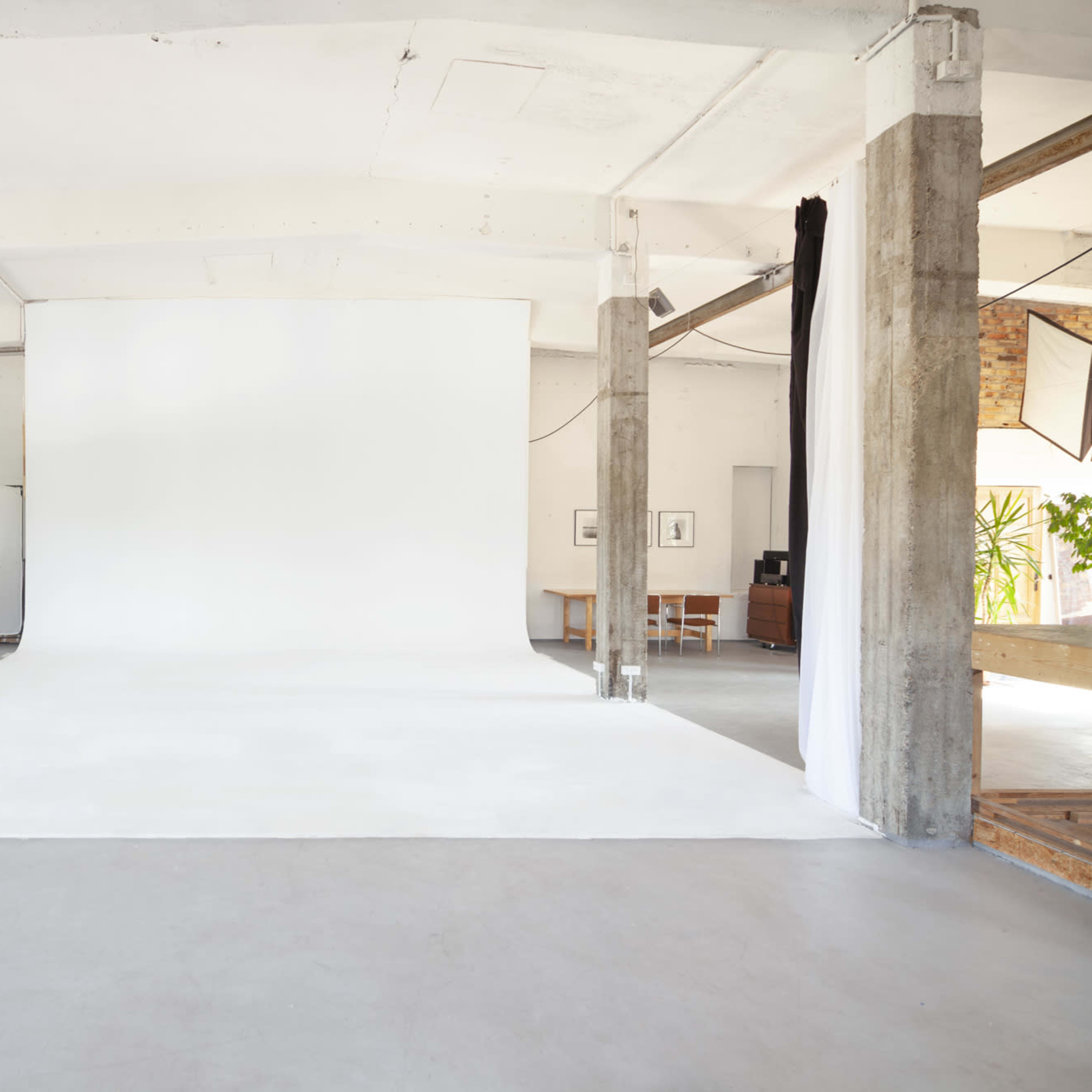 A spacious photography studio with a white backdrop and concrete pillars, featuring a minimalist design and neutral colors.