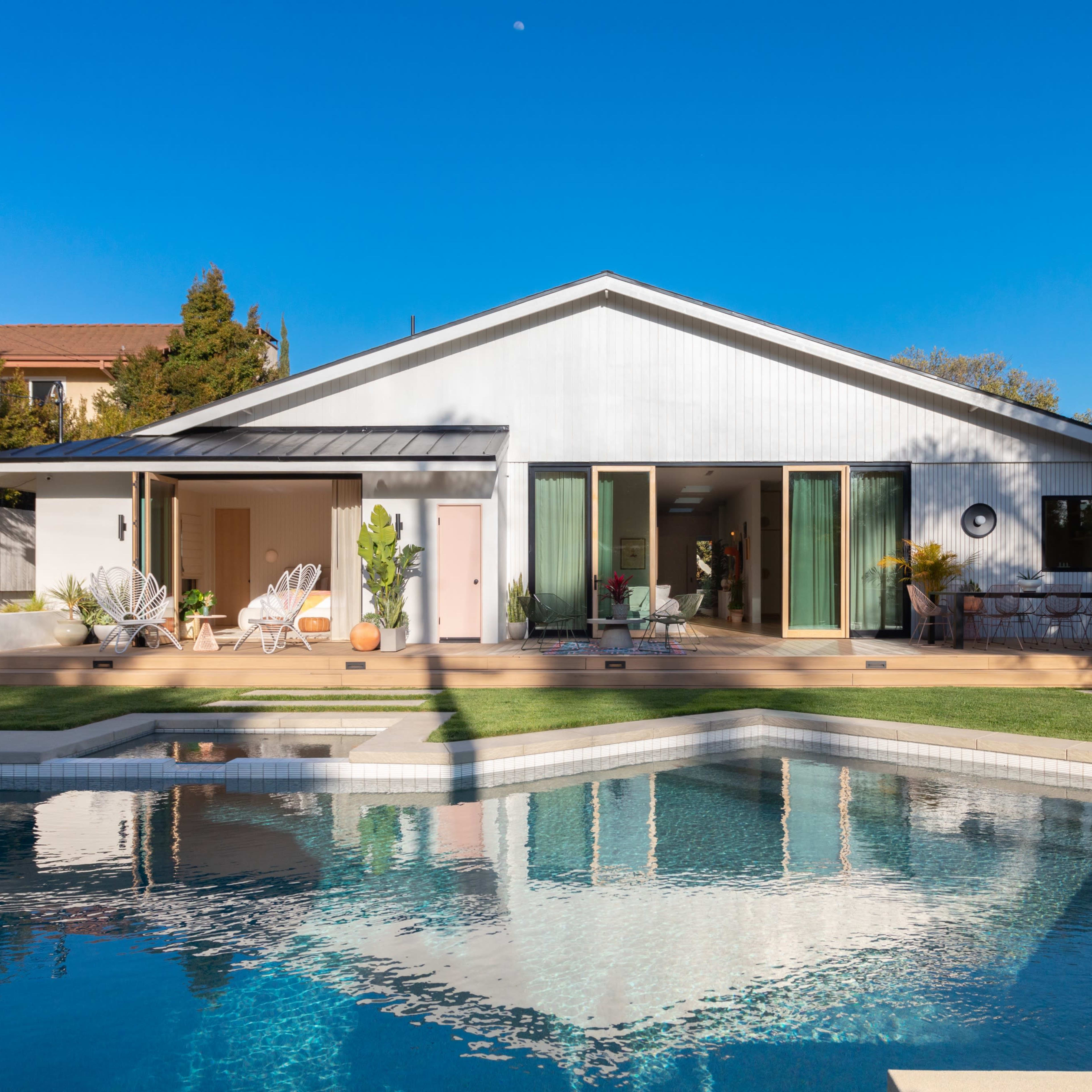 A modern house with large windows and a deck overlooks a swimming pool surrounded by grass and plants.