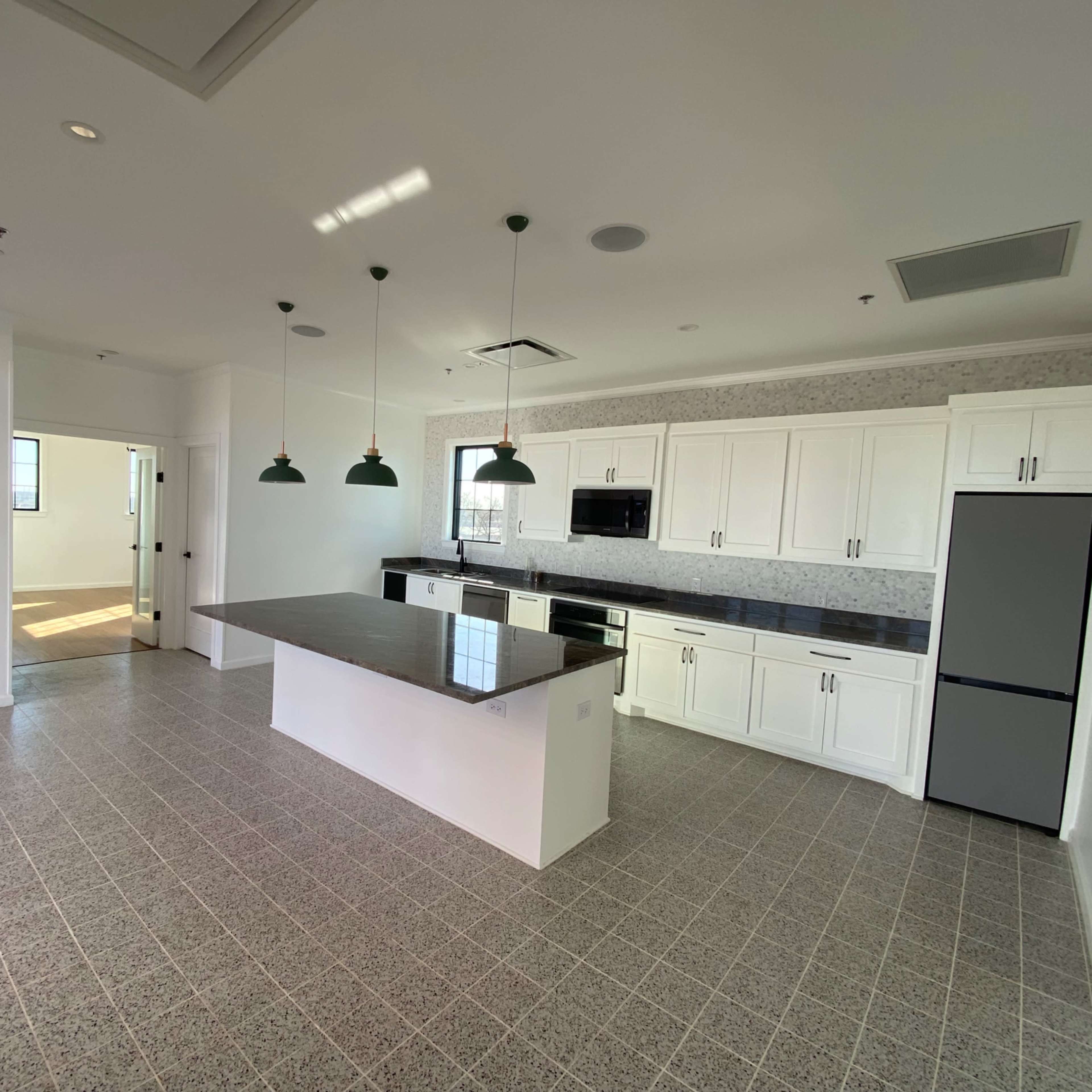 A modern kitchen with white cabinets, a dark countertop, and pendant lights, adjacent to a connected open space.