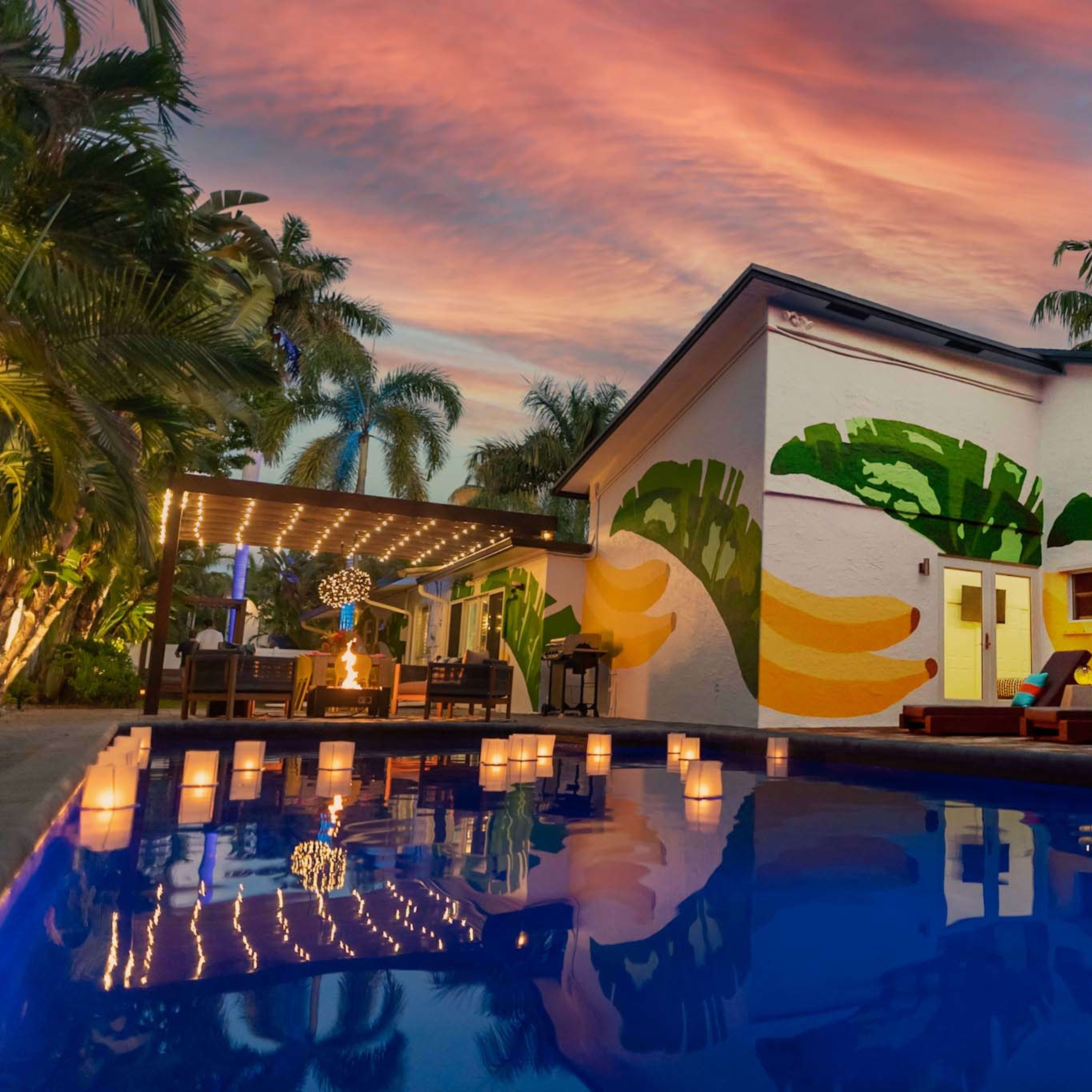 A modern house with a colorful mural and a swimming pool reflects a vibrant sunset, surrounded by palm trees and outdoor seating.