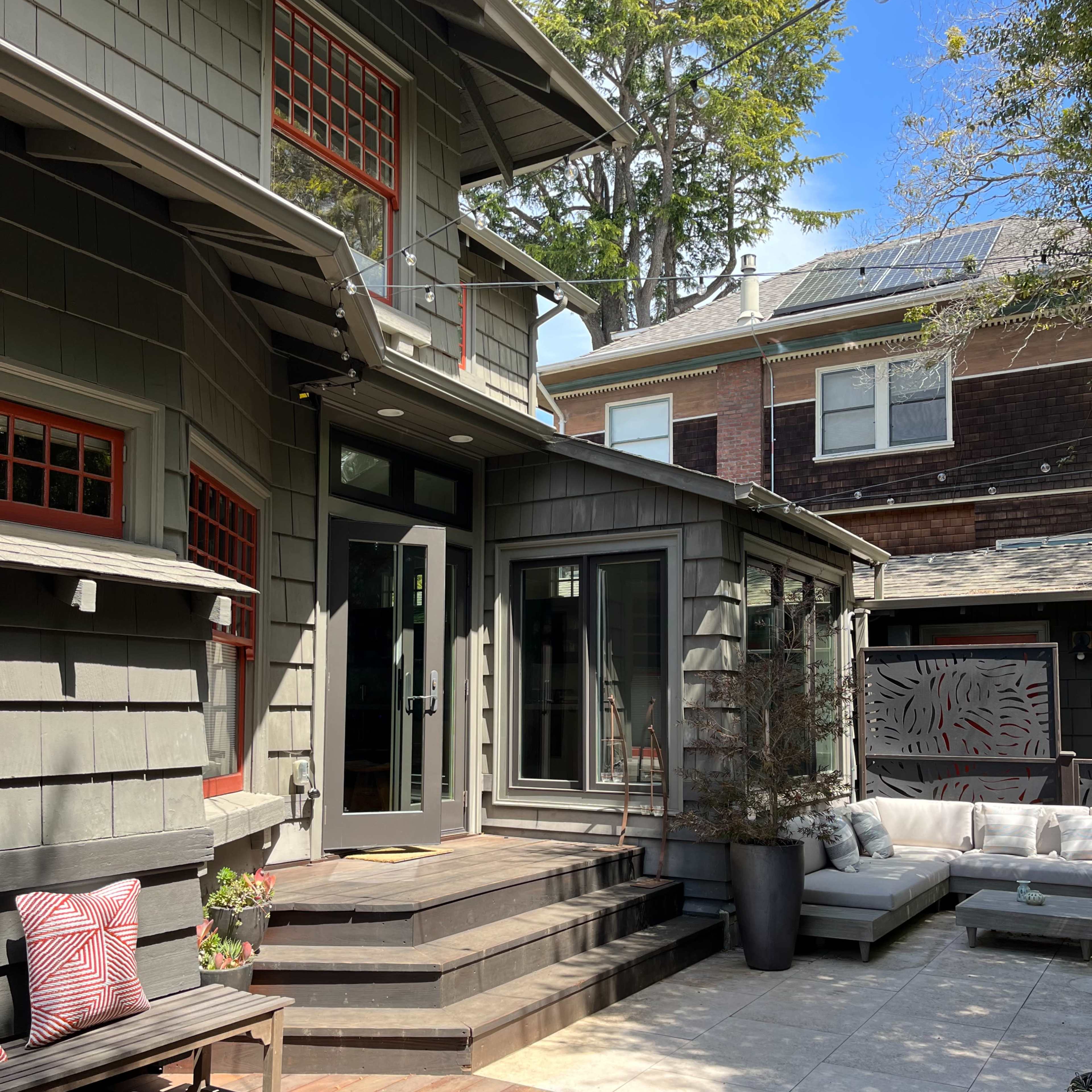 A modern backyard patio area featuring a wooden deck, steps leading to a gray house with red accents, and outdoor seating arrangements.