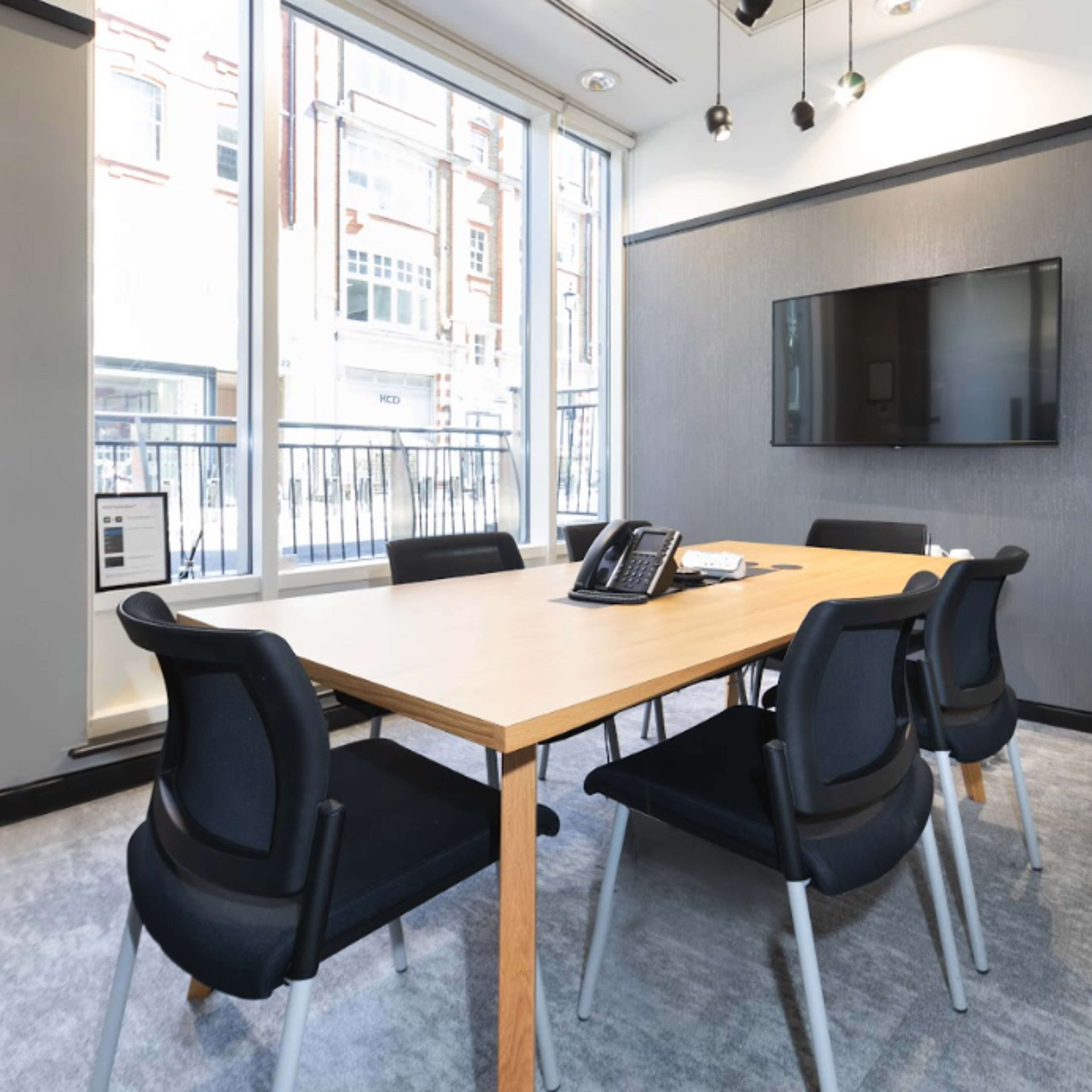 A modern conference room with a wooden table surrounded by black chairs, a wall-mounted TV, and large windows letting in natural light.