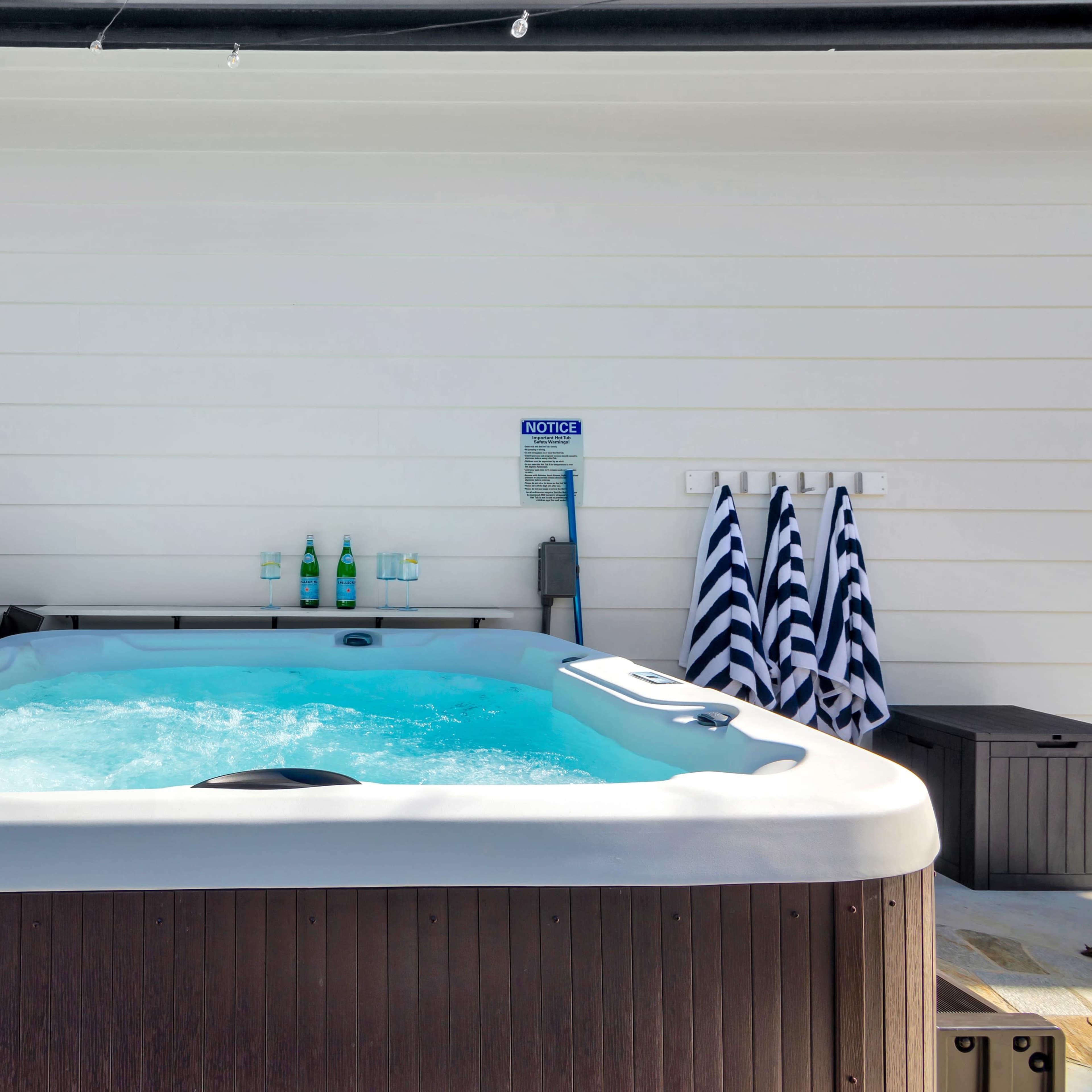 A hot tub filled with bubbling water is positioned on a wooden deck against a white wall, with towels and cleaning supplies nearby.