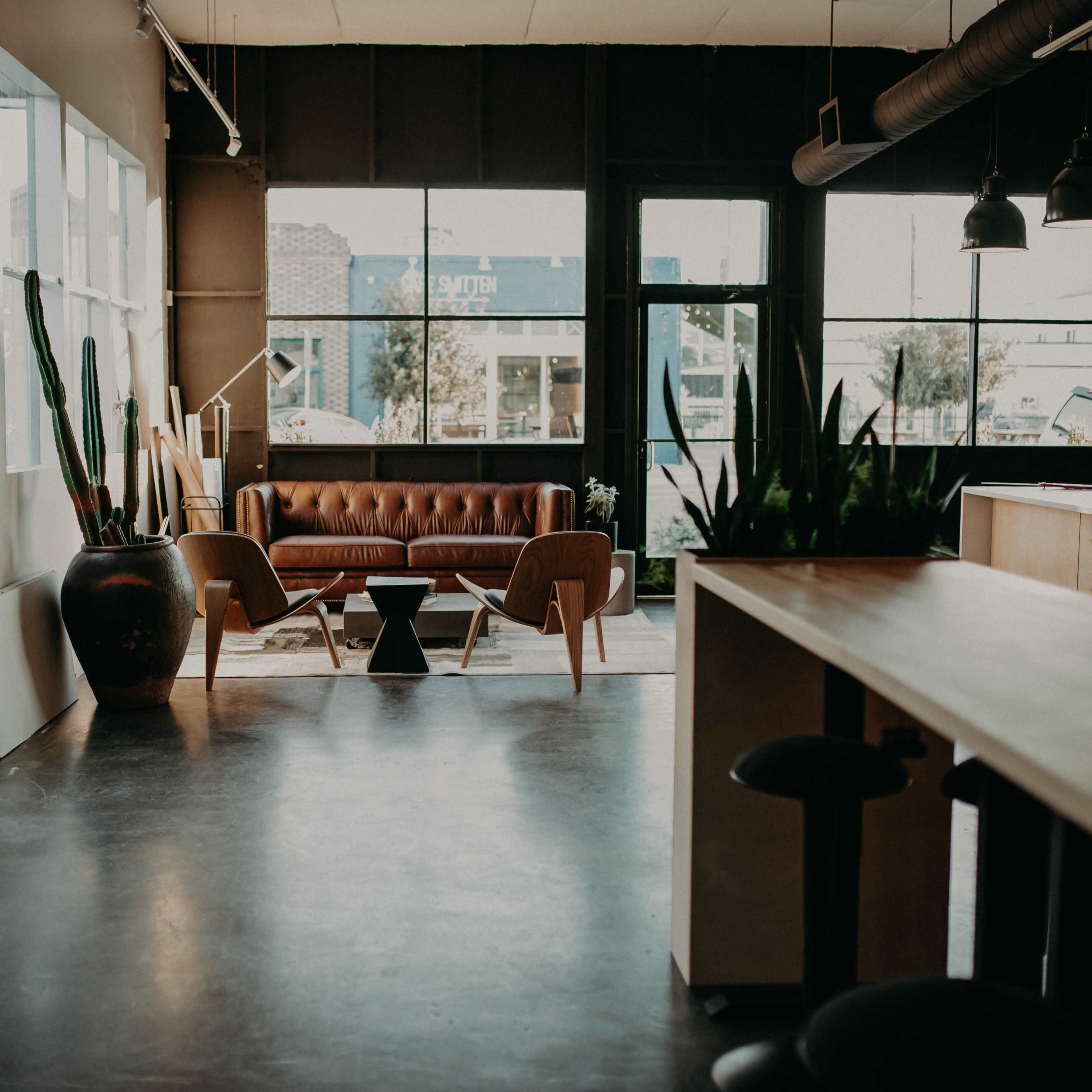 A modern interior space featuring a brown leather couch, wooden furniture, large windows, and various plants.