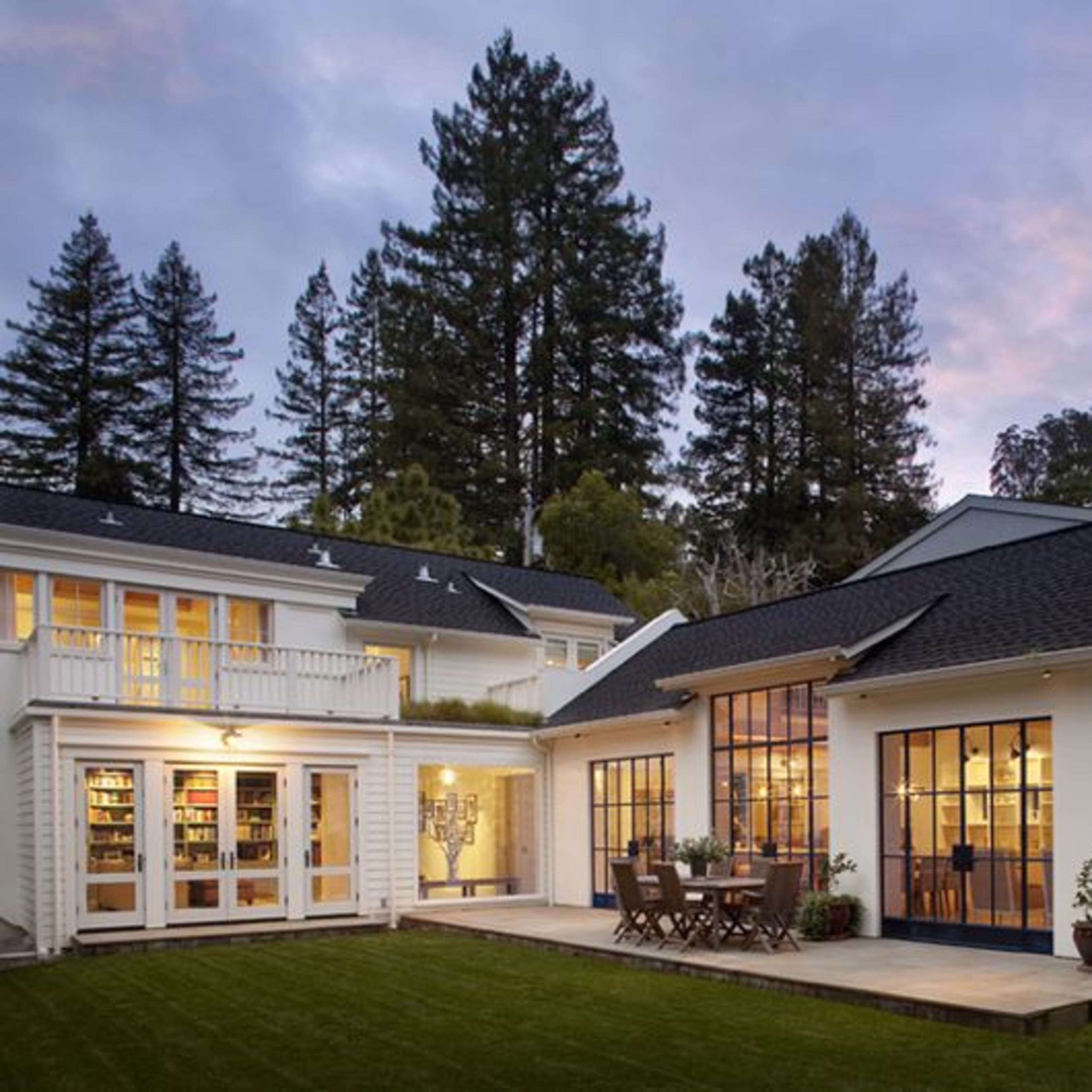 A modern two-story house with large windows, a stone chimney, and a wooden patio surrounded by trees.