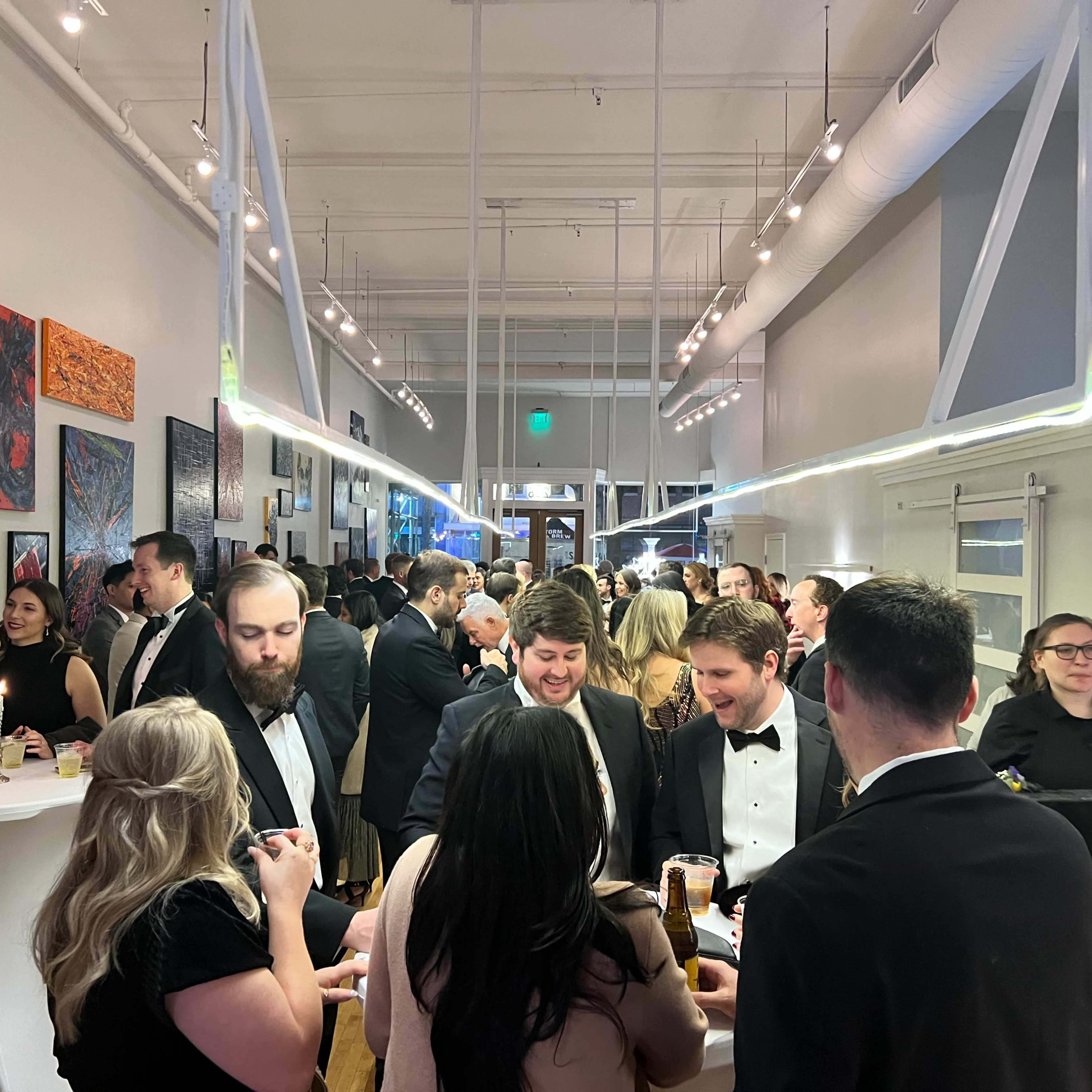 A crowded indoor venue featuring guests in formal attire engaged in conversation and socializing amidst colorful artwork on the walls.