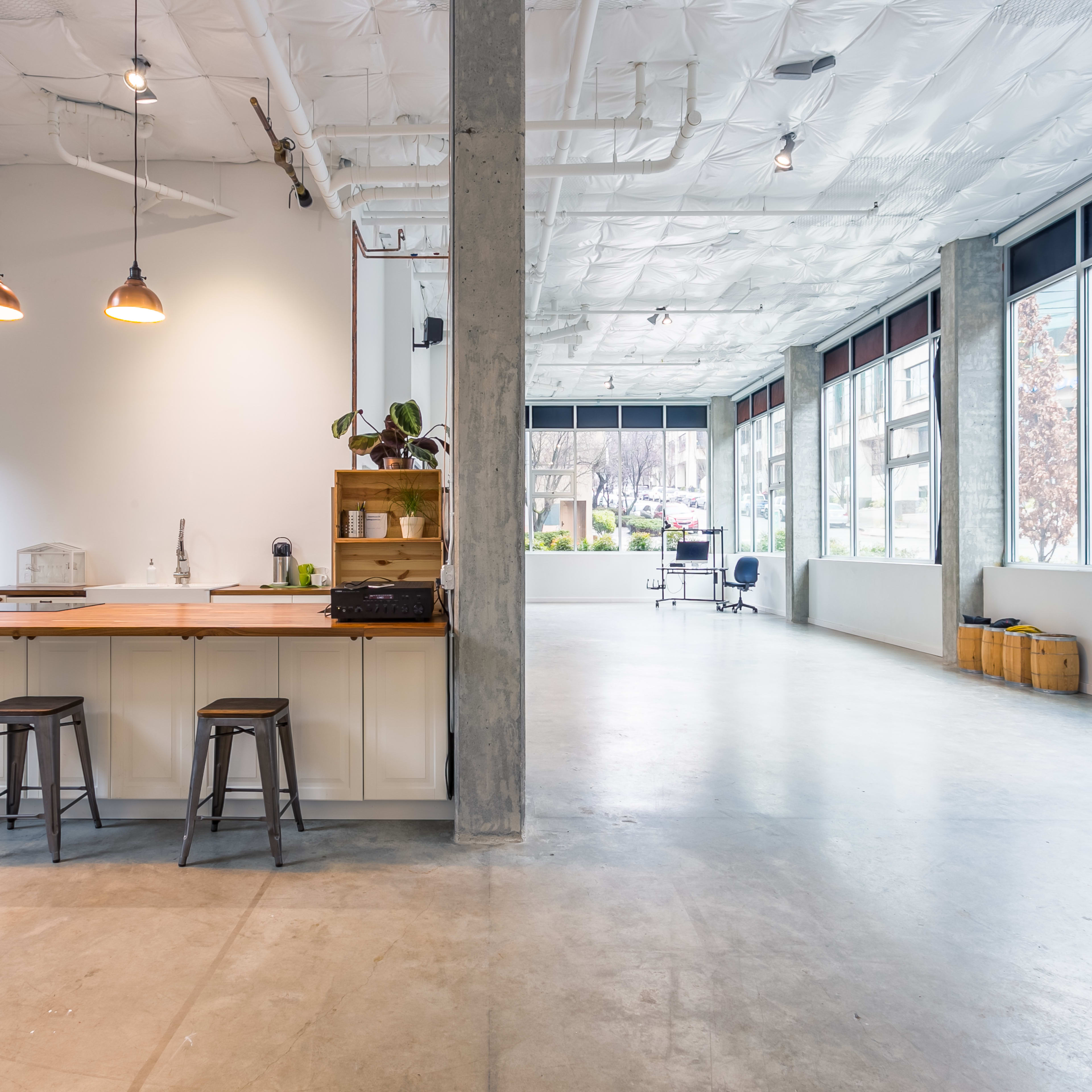 An airy, modern workspace with a kitchen island, bar stools, large windows, and a polished concrete floor.