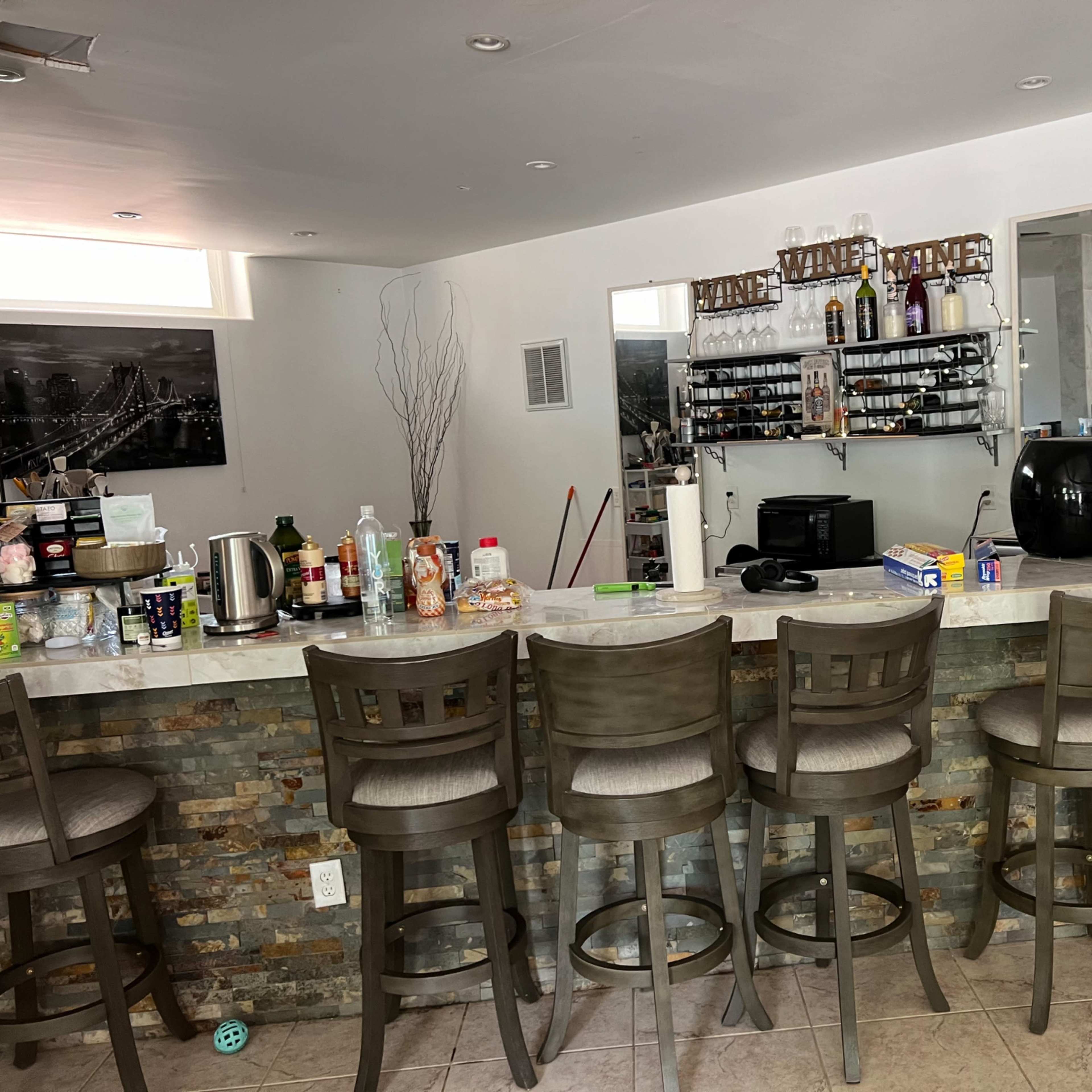 A modern home bar features several high stools around a stone-topped counter, with shelves of drinks and appliances in the background.