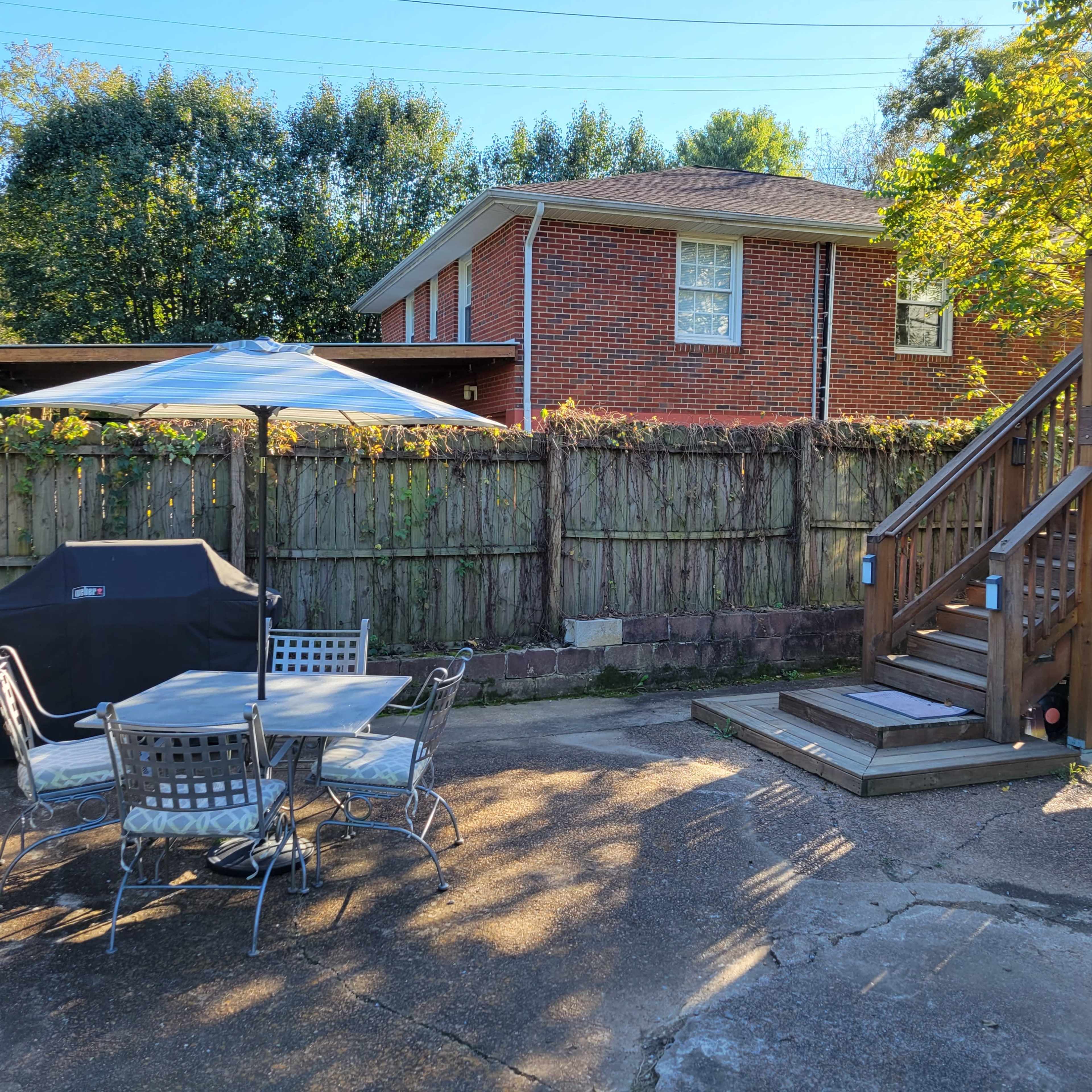 A backyard features a seating area with a table and chairs, a grill under an umbrella, and a wooden deck leading to a house.