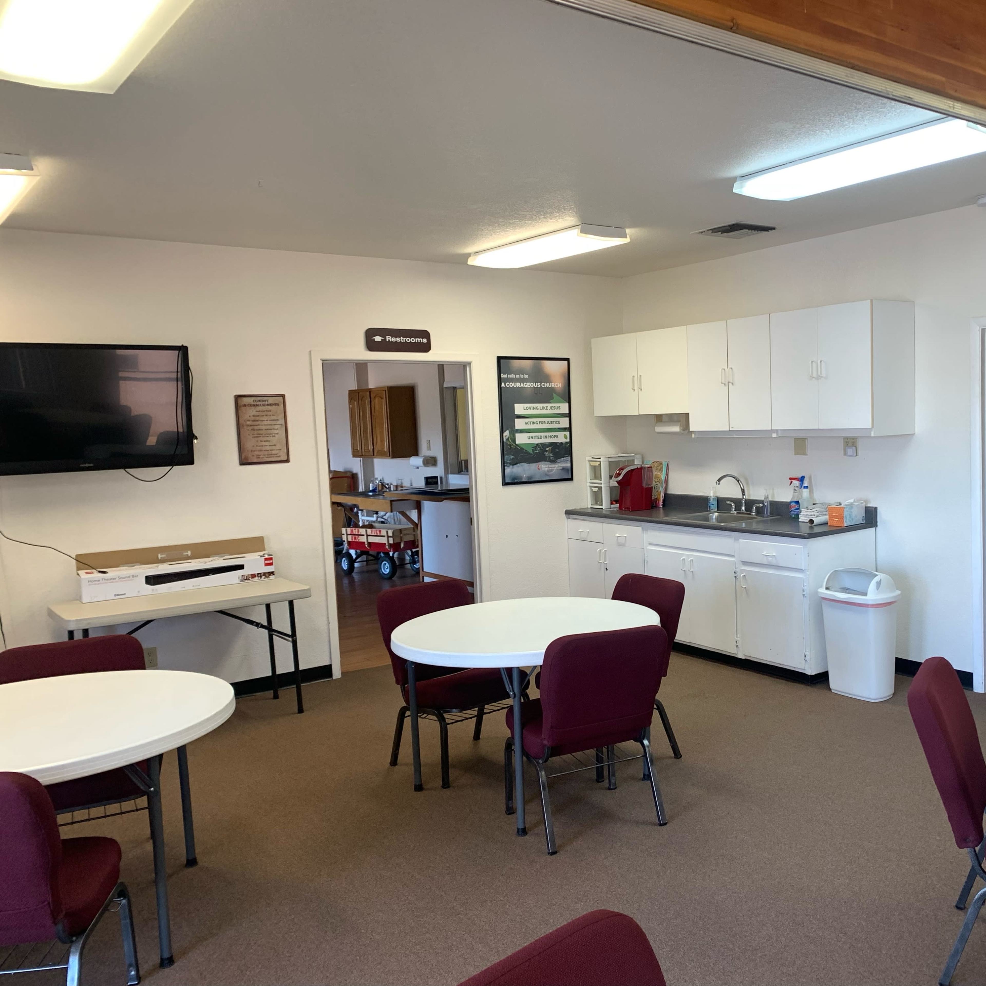 A clean, well-lit communal kitchen and dining area featuring several round tables with chairs, a sink, and a television on the wall.