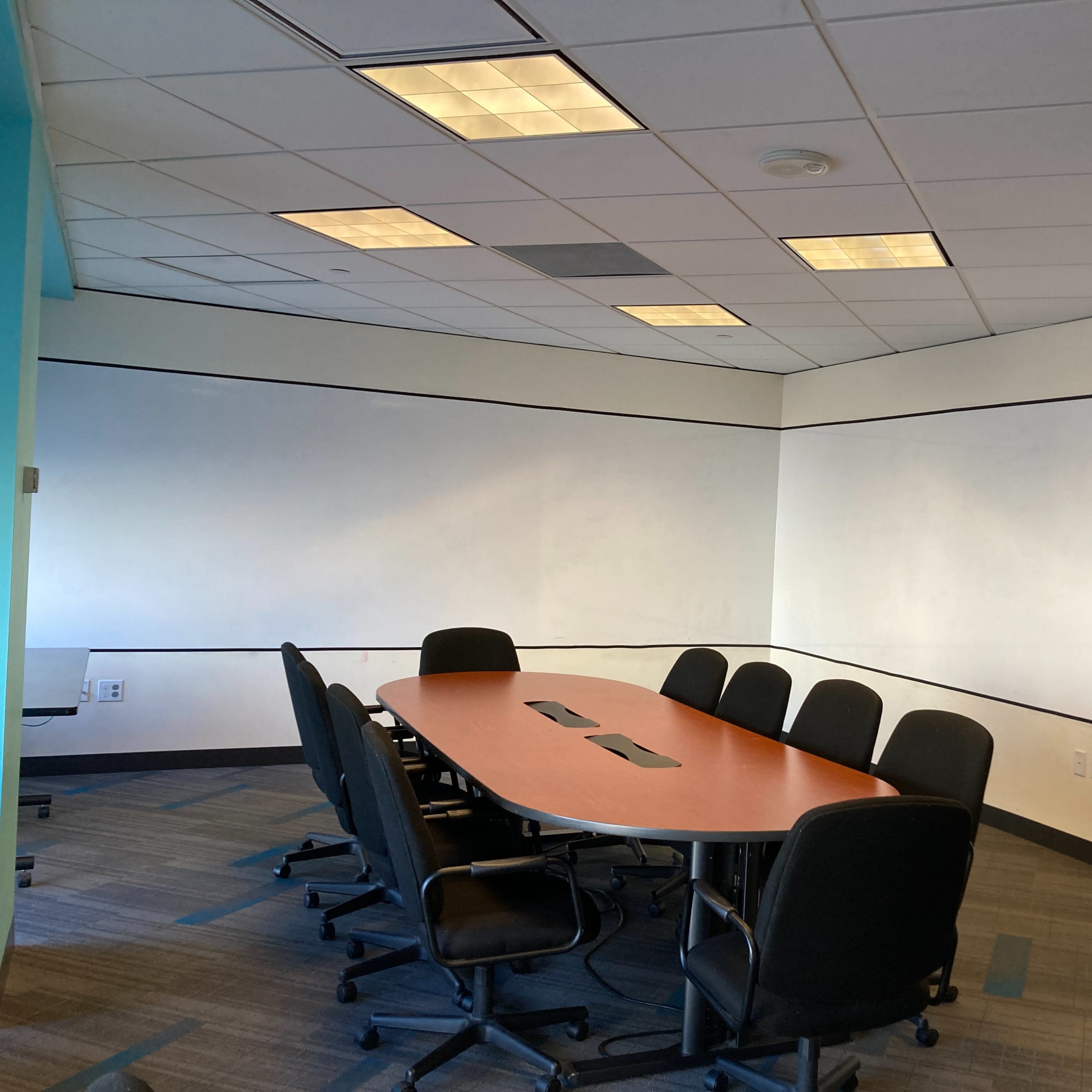 A conference room features a large rectangular table surrounded by chairs, with white walls and fluorescent lighting.