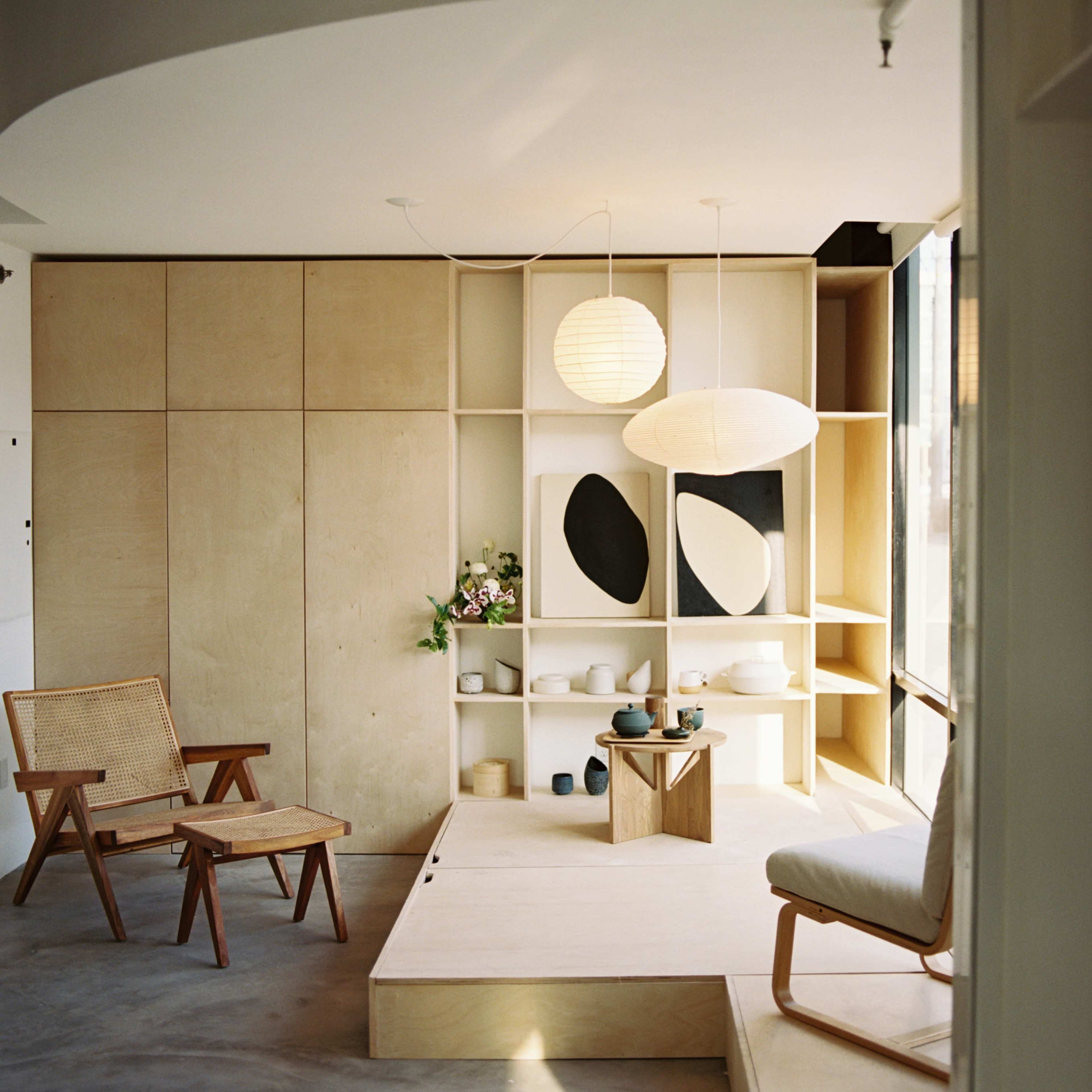 A minimalist interior space featuring a wooden platform, a lounge chair, and a light fixture above a display of artistic decor and pottery.