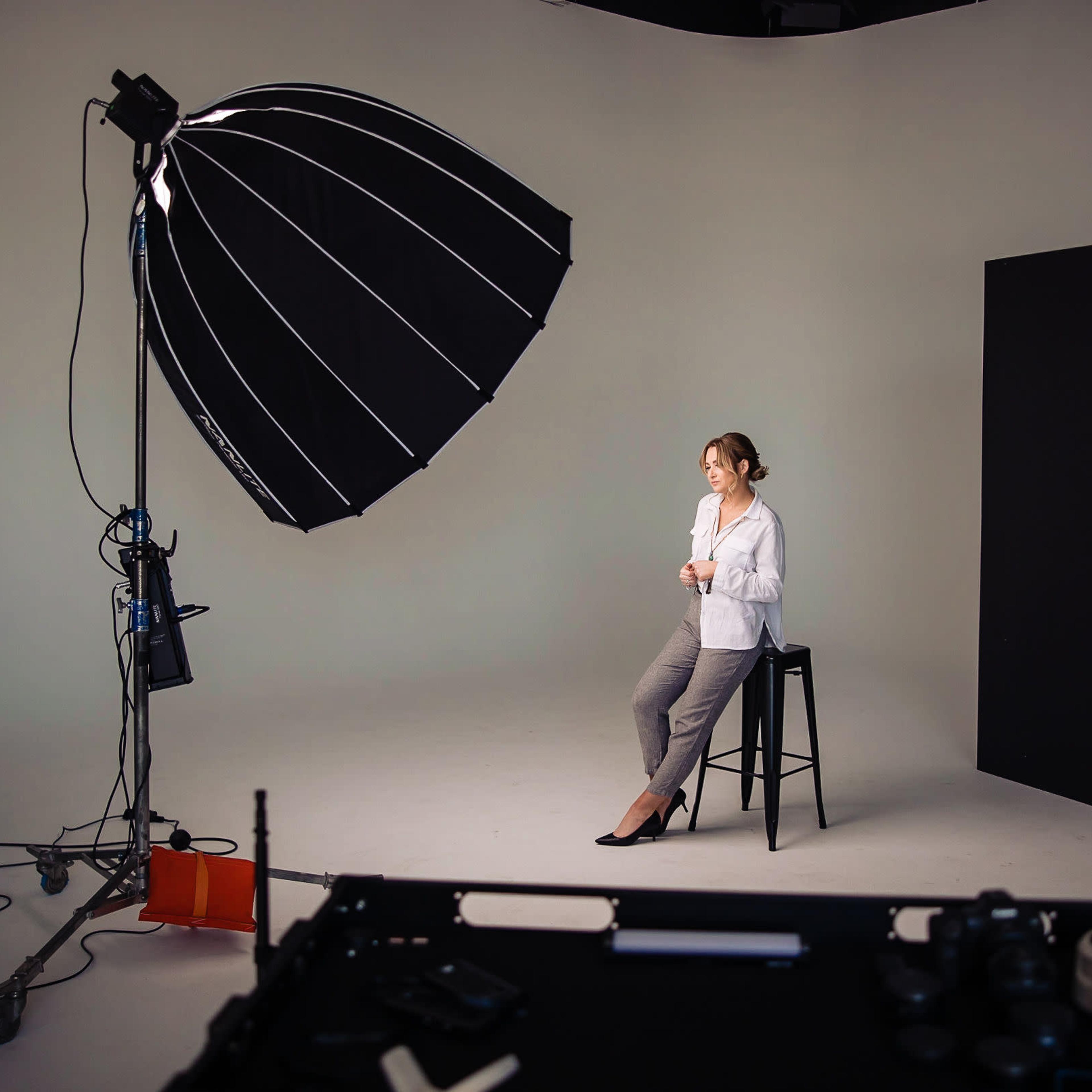 A woman sits on a stool in a photography studio, surrounded by lighting equipment and a black backdrop.