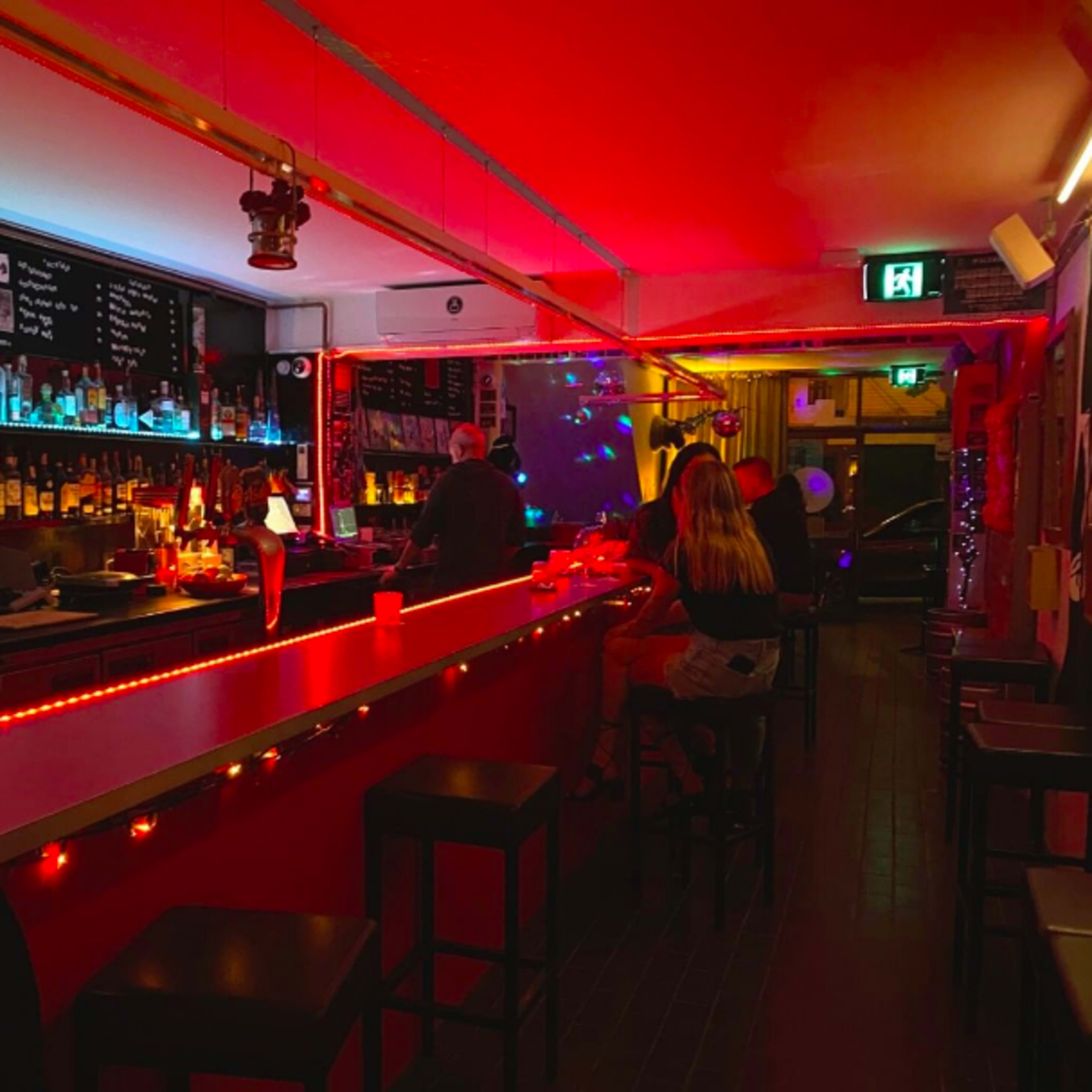 A dimly lit bar with red and blue lighting, featuring a long bar counter and patrons sitting on stools.
