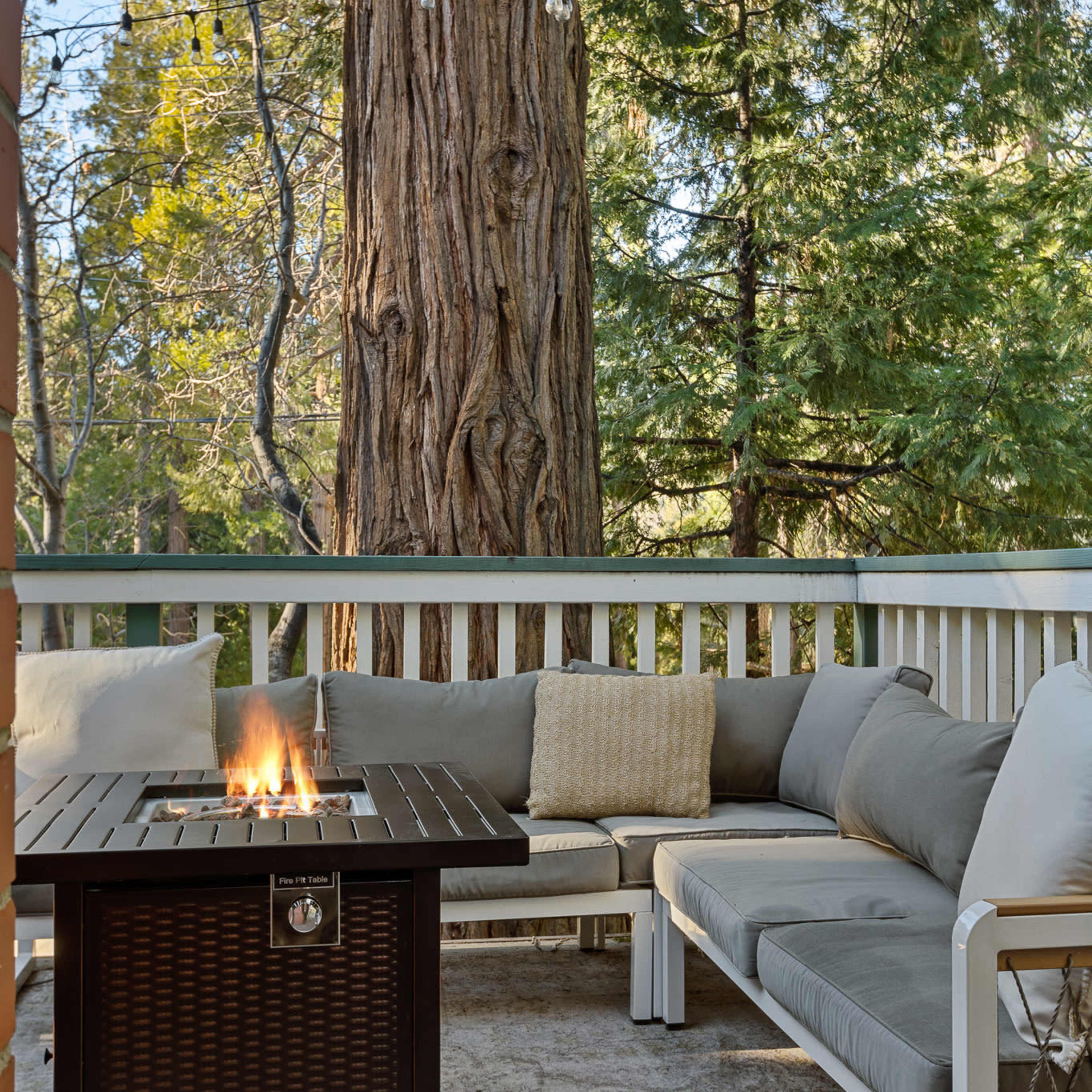 A cozy outdoor seating area features a fire table surrounded by a sectional sofa on a patio, with tall trees in the background.