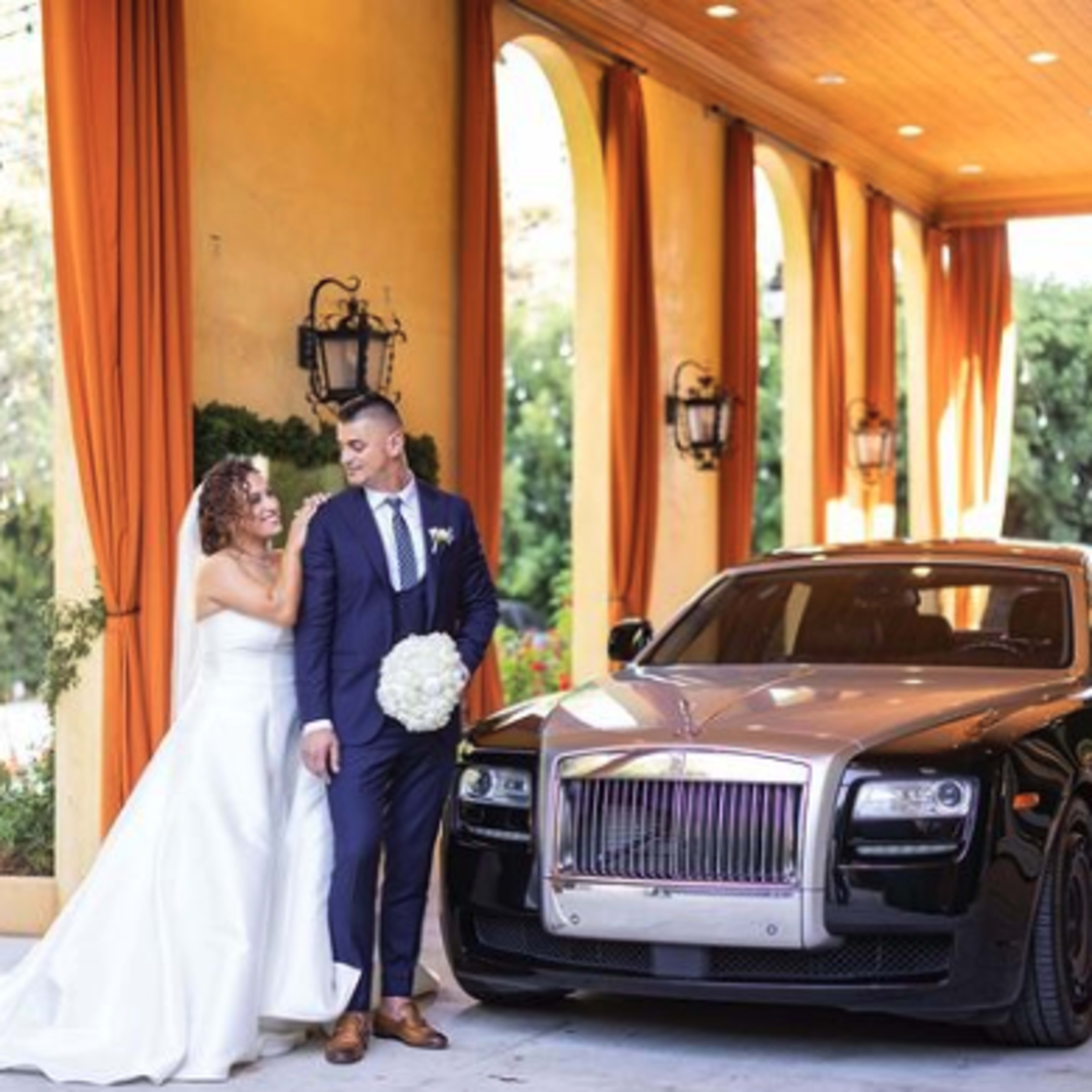 A bride in a white gown and a groom in a dark suit stand beside a luxury black Rolls-Royce in a patio setting.