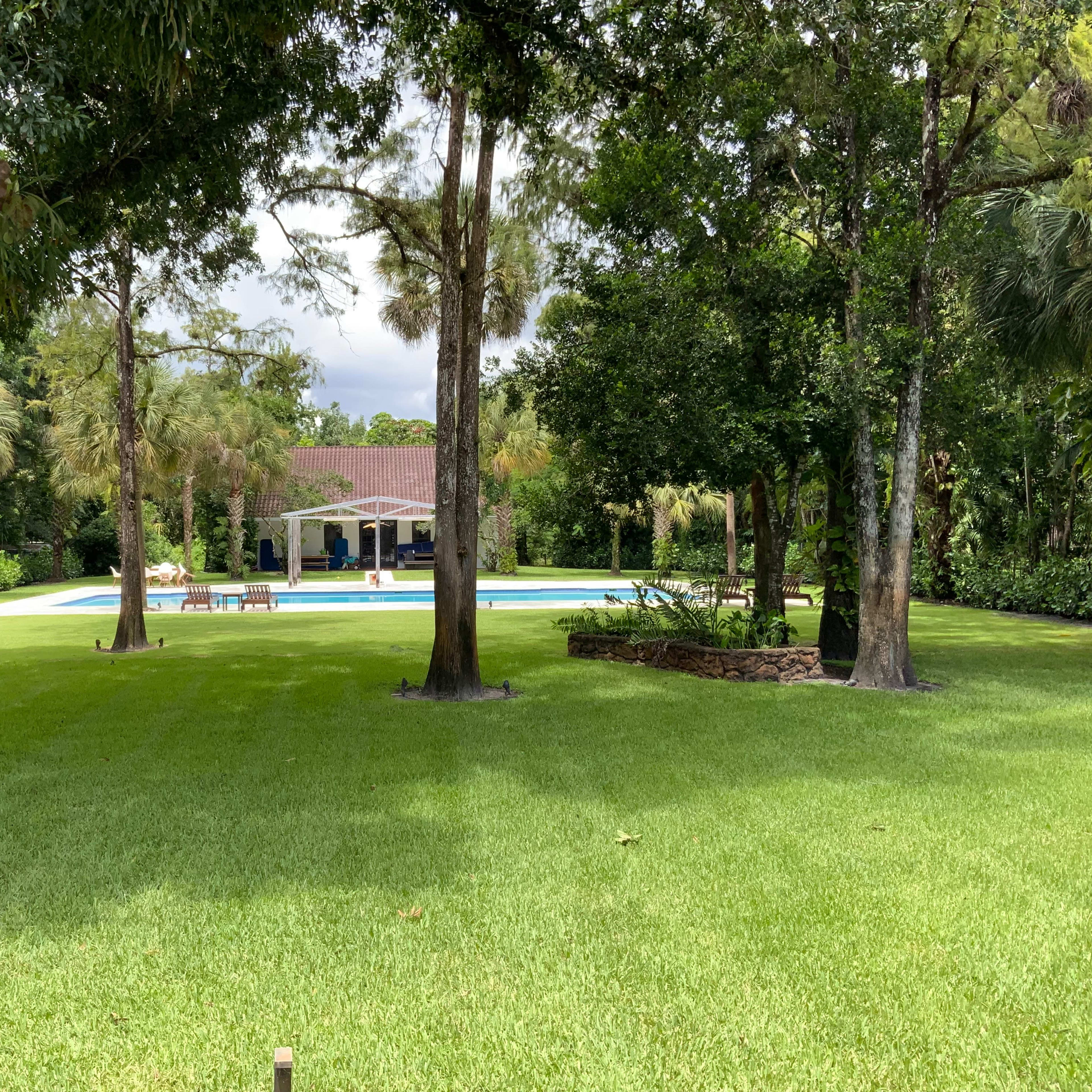 A spacious backyard with a swimming pool surrounded by lush green grass and trees, featuring a house in the background.