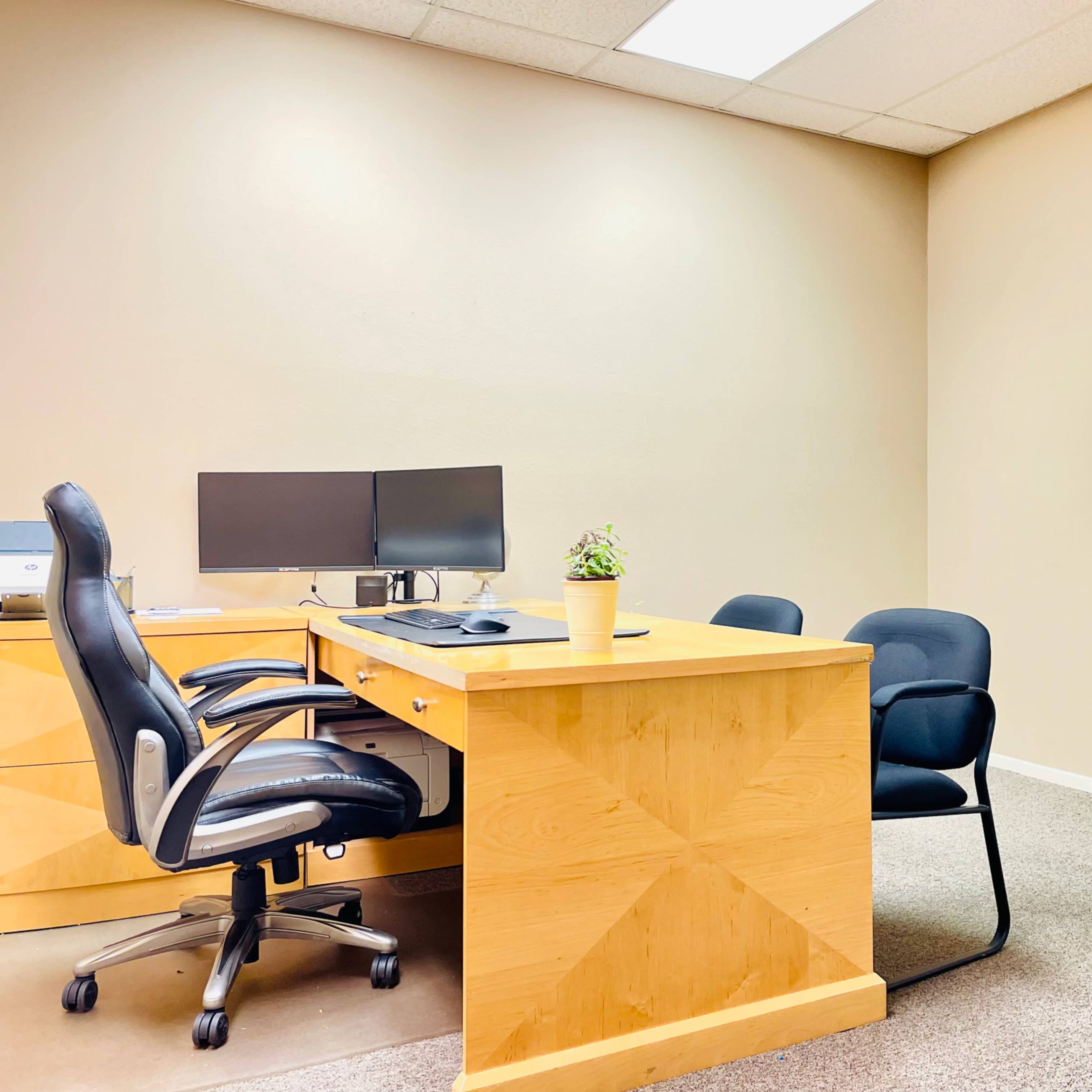 A simple office setup featuring a wooden desk with two computer monitors, an office chair, a potted plant, and a whiteboard on the wall.