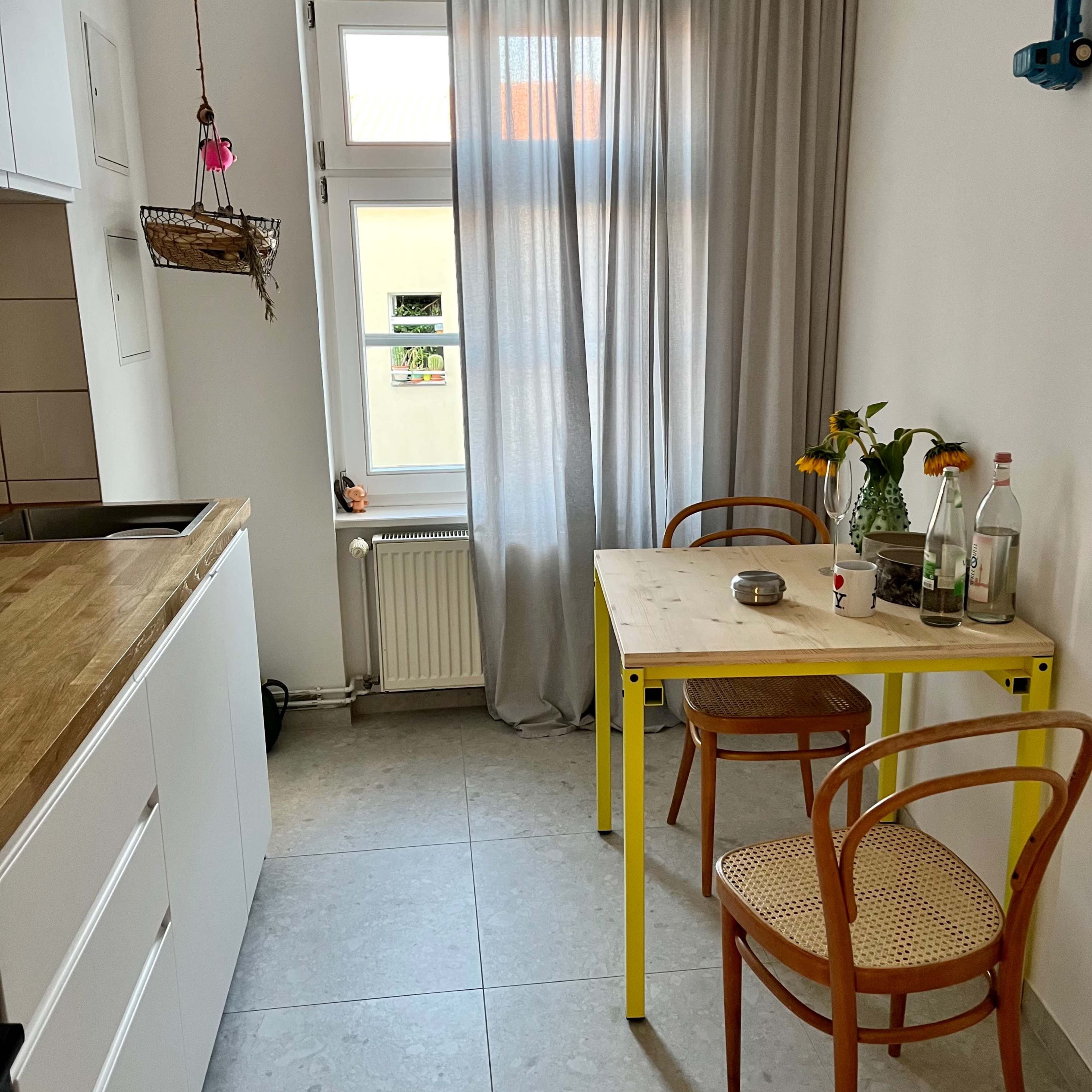 A small kitchen features a wooden counter, a table with yellow legs, two chairs, a vase of flowers, and a window with sheer curtains.
