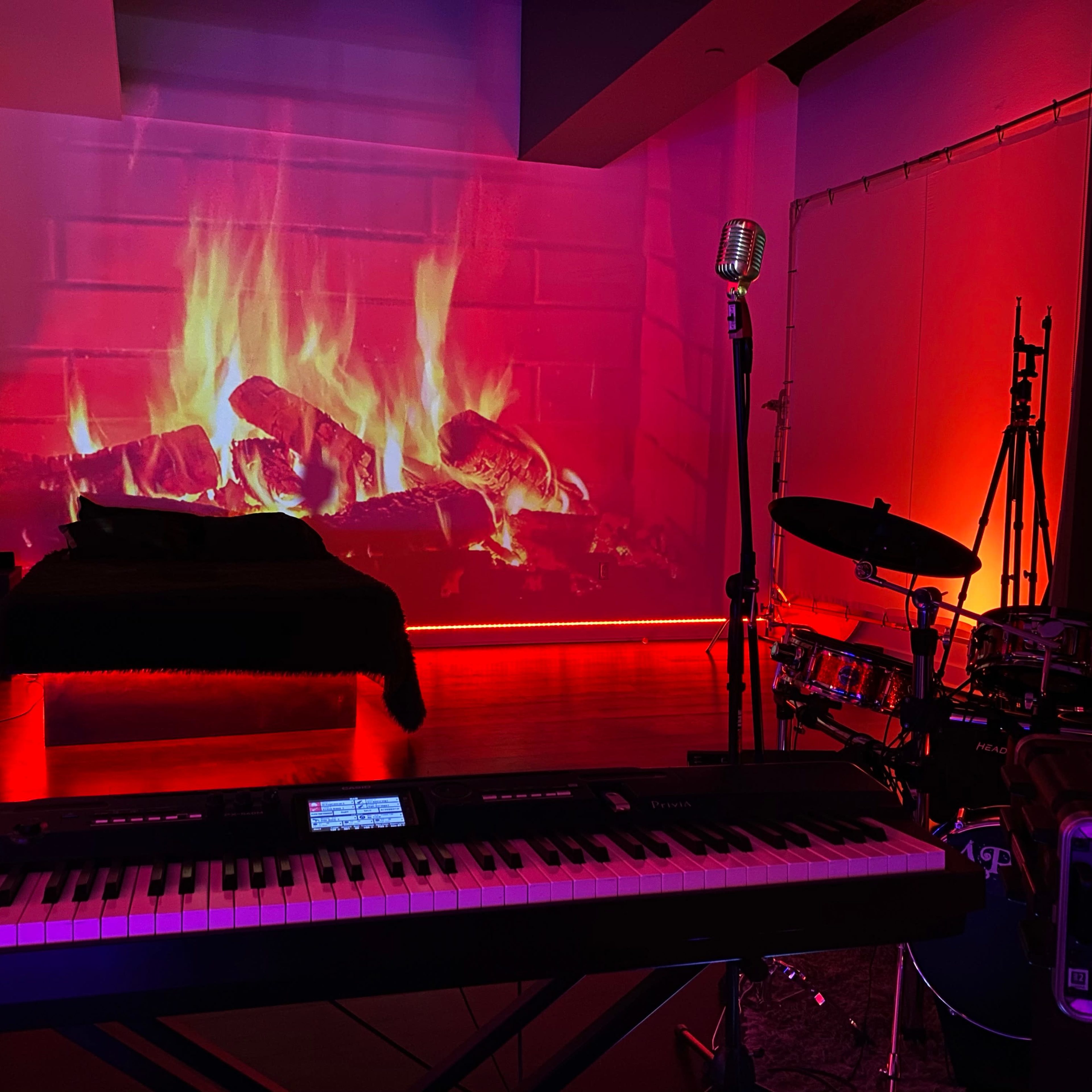 A room features a keyboard, drum set, and a large screen displaying a digitally created fireplace with glowing flames.
