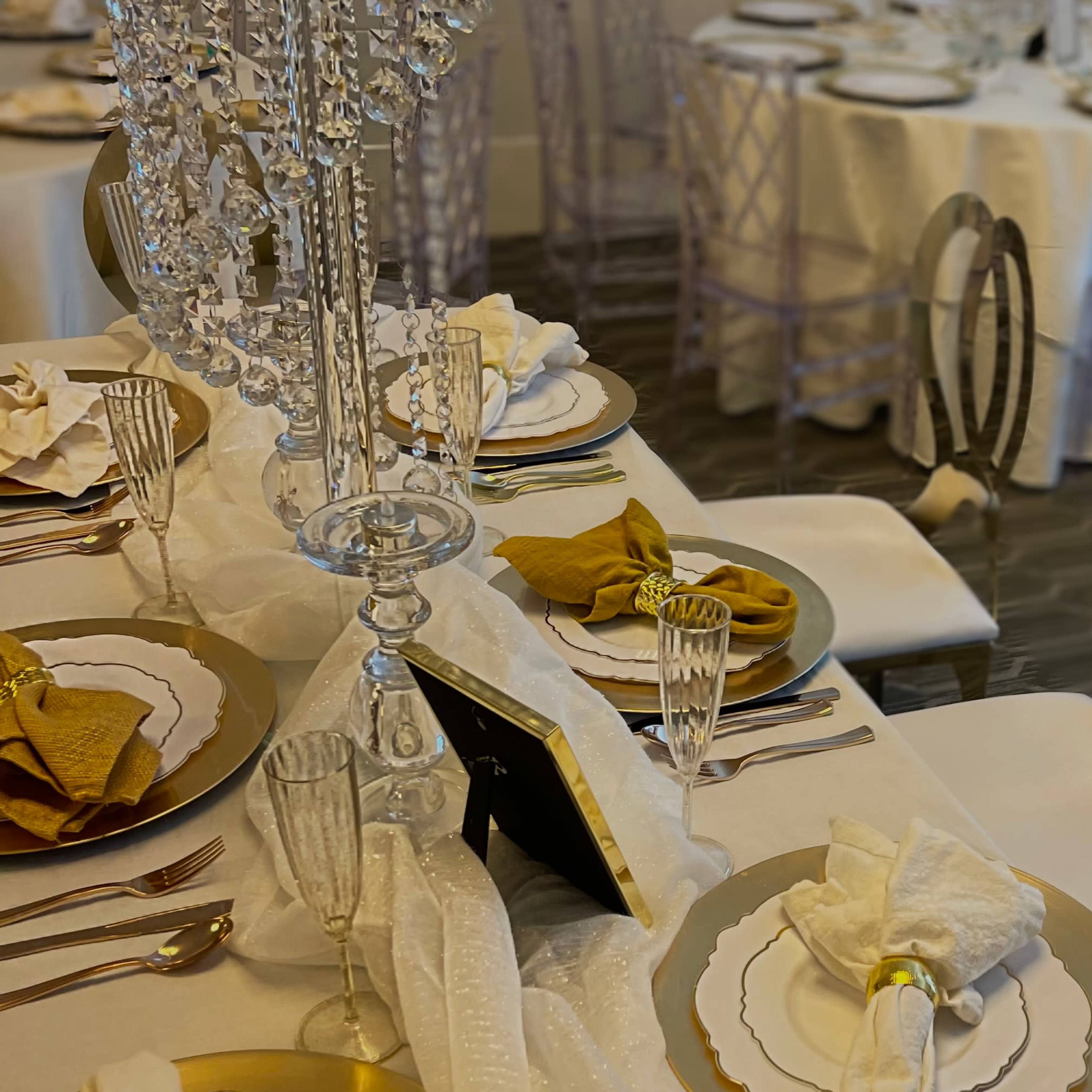 A white table is elegantly set with gold-trimmed plates, crystal glassware, and a decorative chandelier centerpiece.