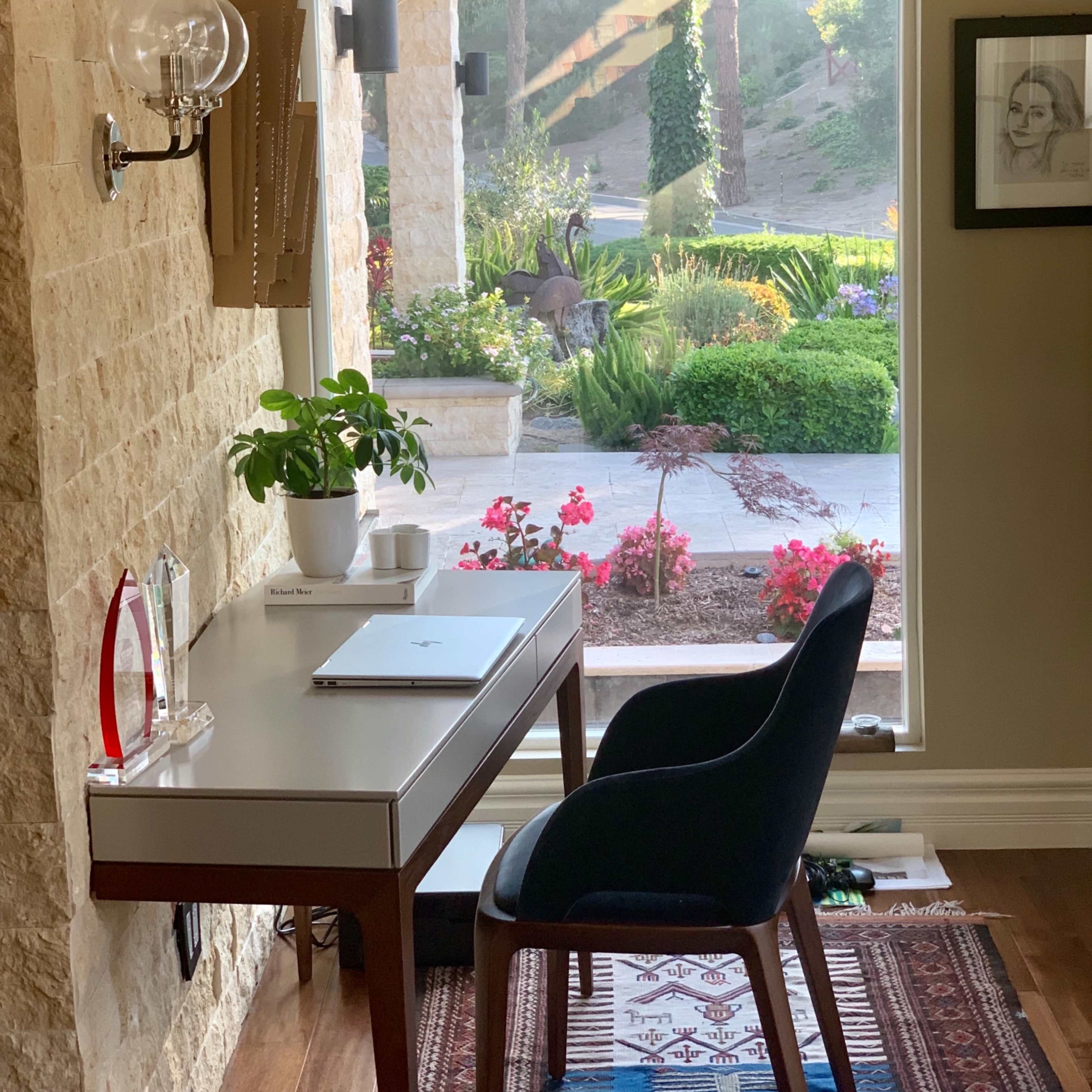 A desk with a plant and a laptop faces a large window overlooking a landscaped garden with flowers and greenery.