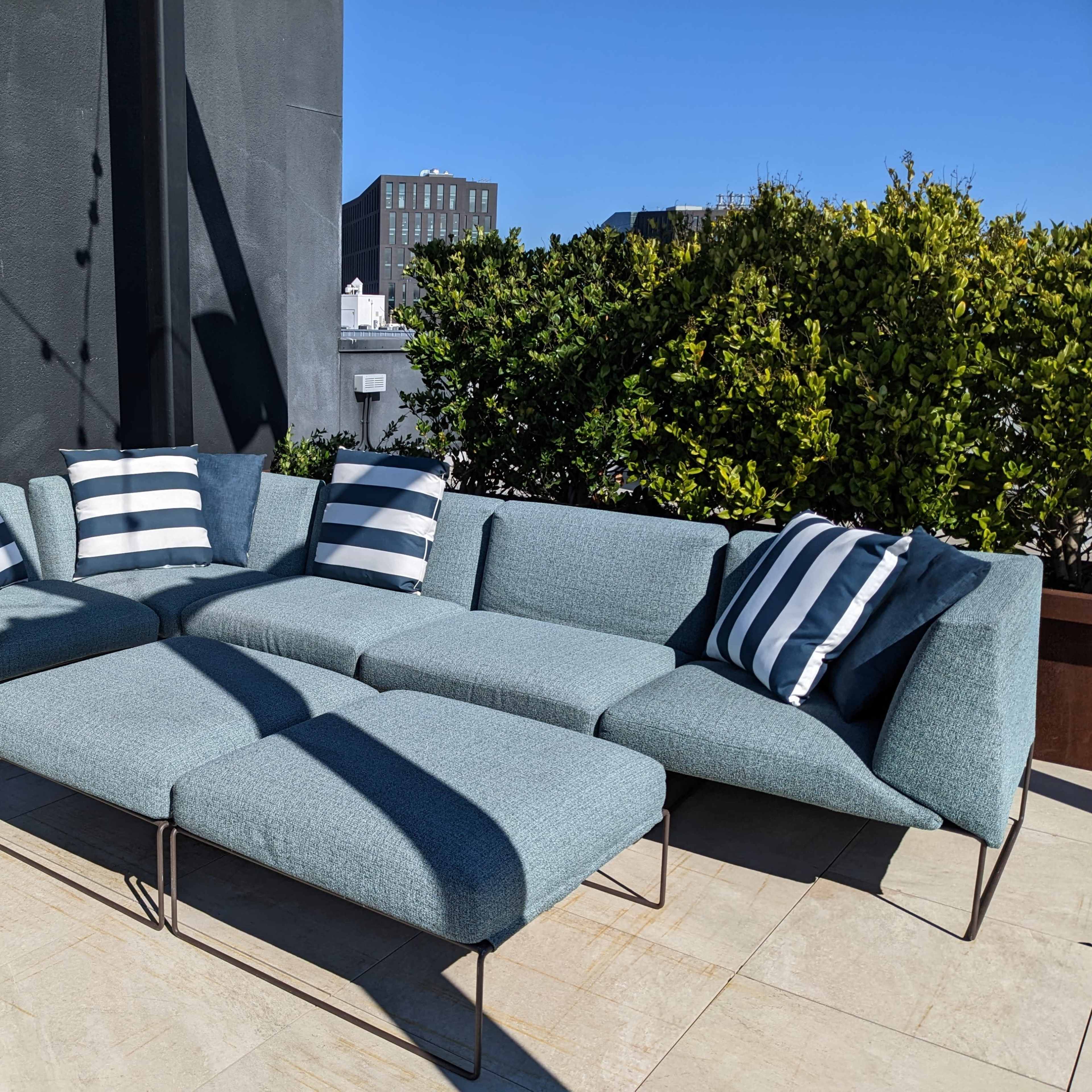 A modern sectional sofa with blue upholstery and striped cushions is positioned on a rooftop terrace surrounded by greenery and a clear blue sky.