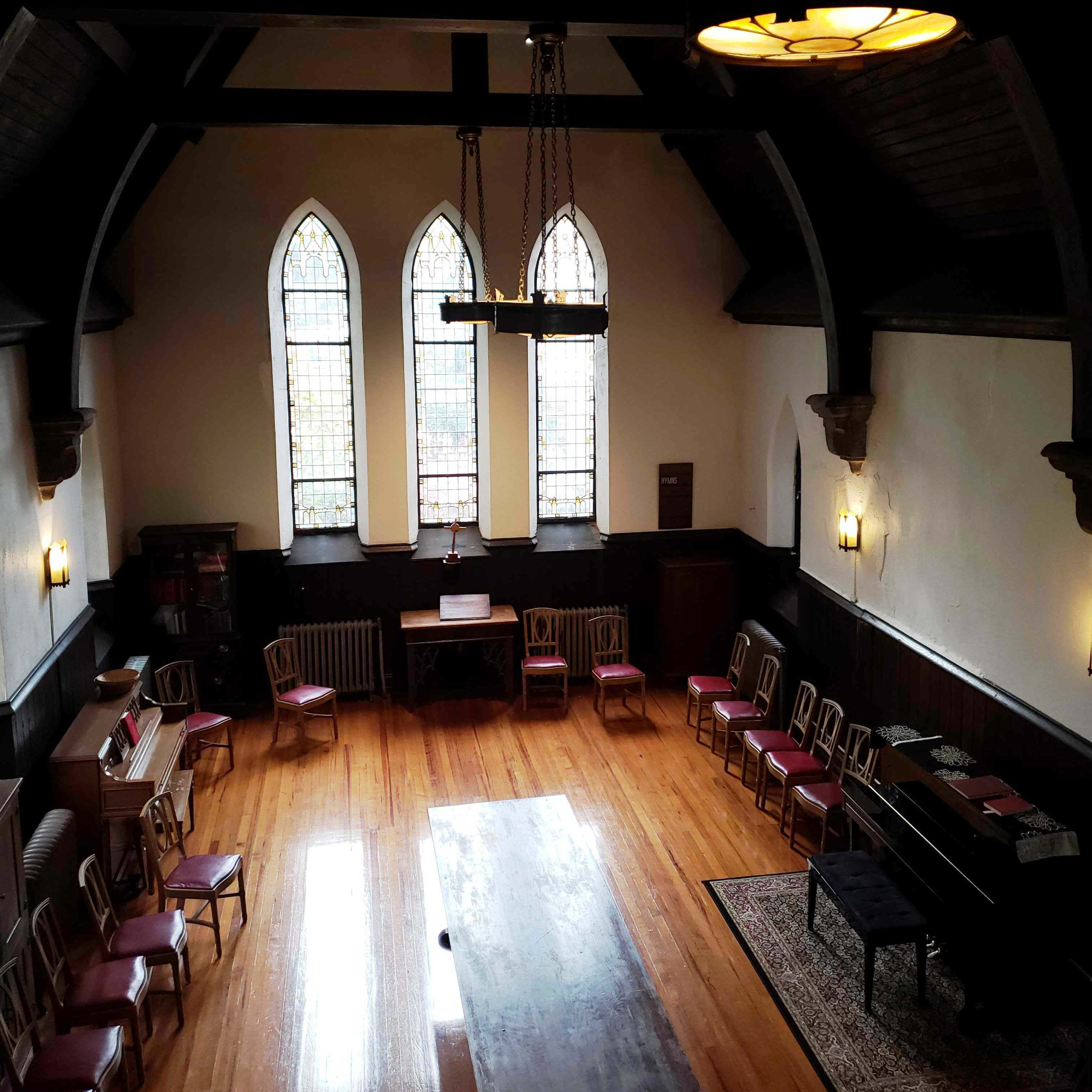An interior room with a long wooden table surrounded by chairs, tall stained glass windows, and wooden beams in the ceiling.
