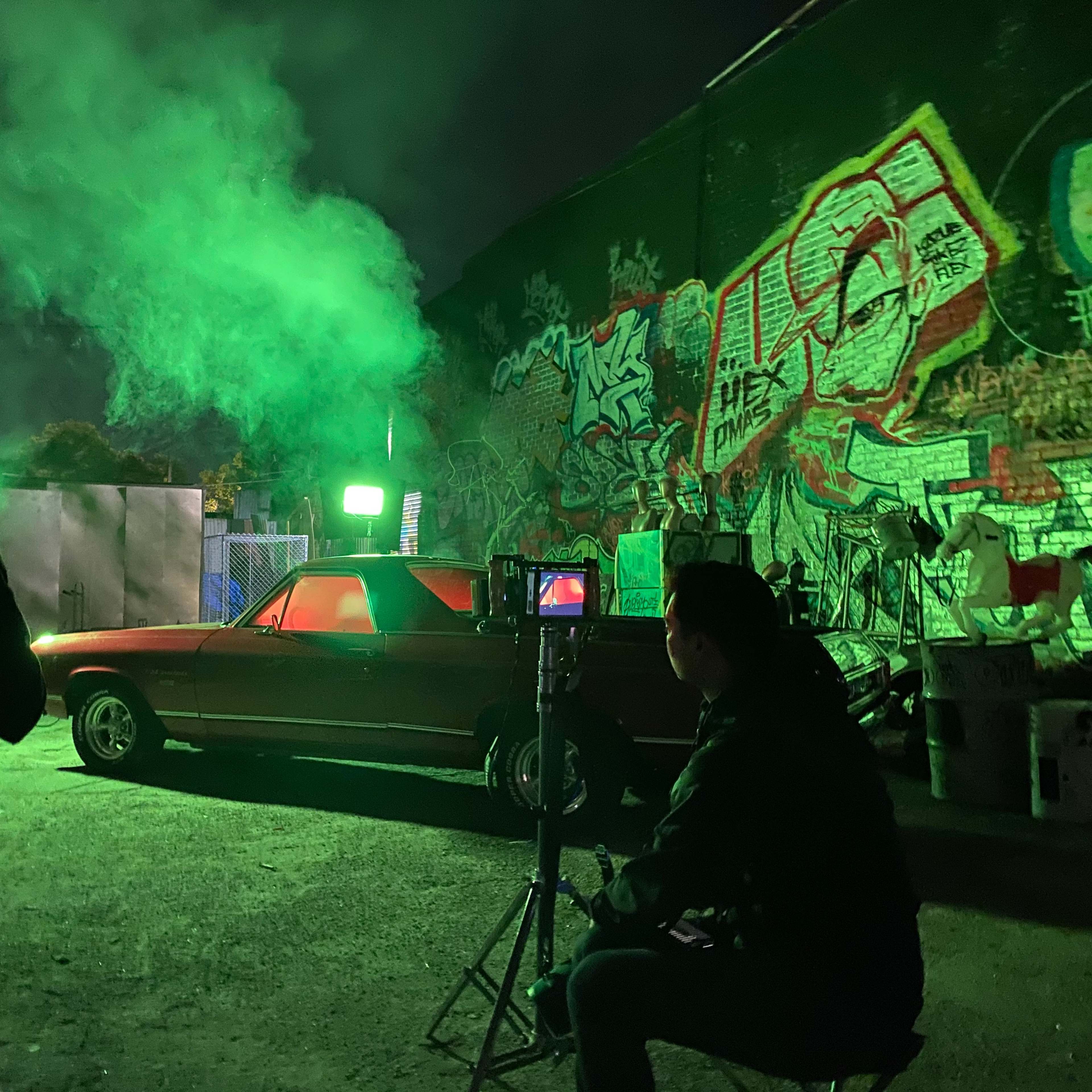 A group of people is filming a scene at night in an alley, featuring a red car illuminated by green lighting against a backdrop of graffiti.