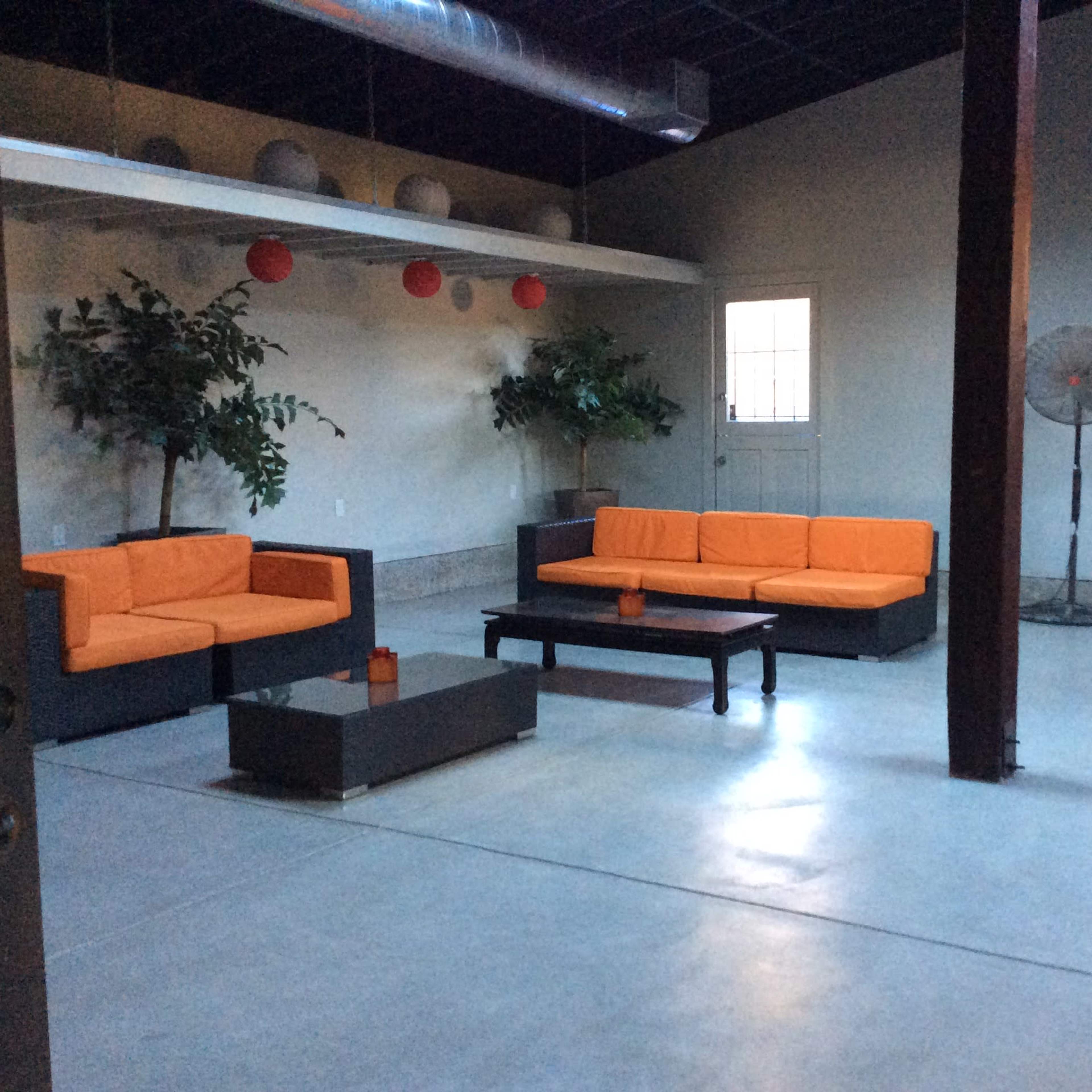 A spacious indoor area with orange sofas, a coffee table, and two large fans near the walls, under a ceiling with hanging decorations.