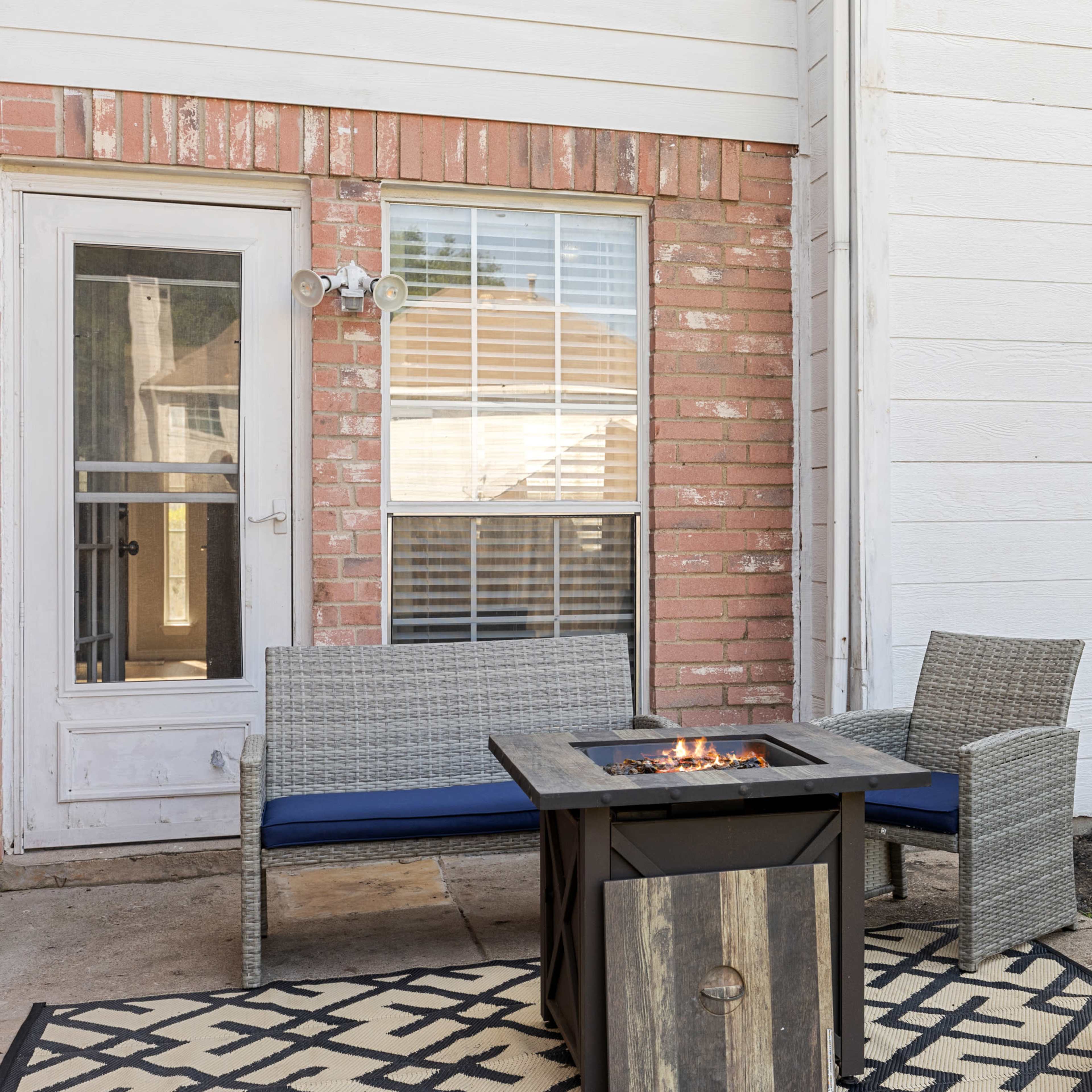A small outdoor patio features gray wicker furniture arranged around a fire pit, adjacent to a brick wall and a sliding glass door.