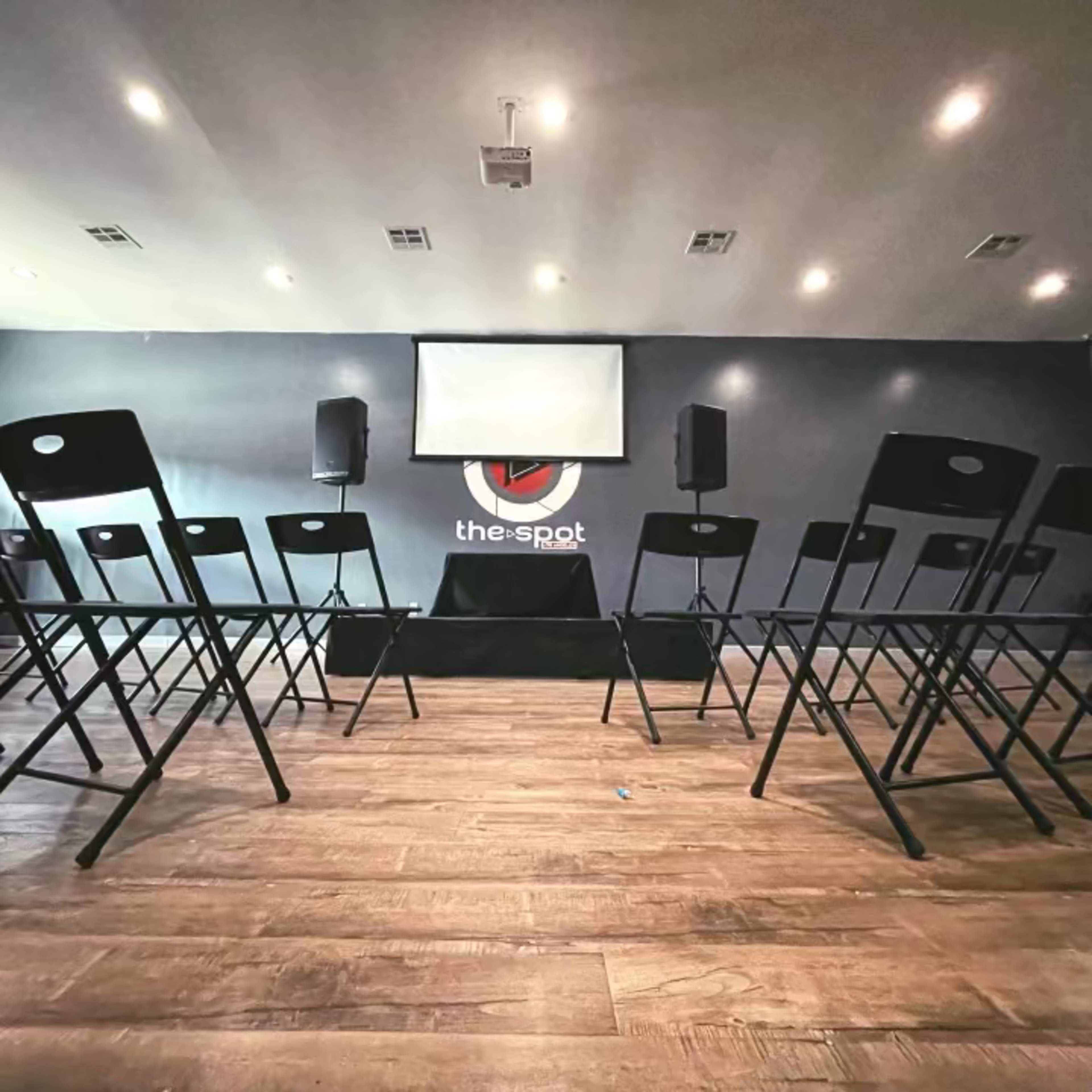 A seating arrangement of black folding chairs facing a blank projection screen in a room with a dark wall labeled "the-spot."